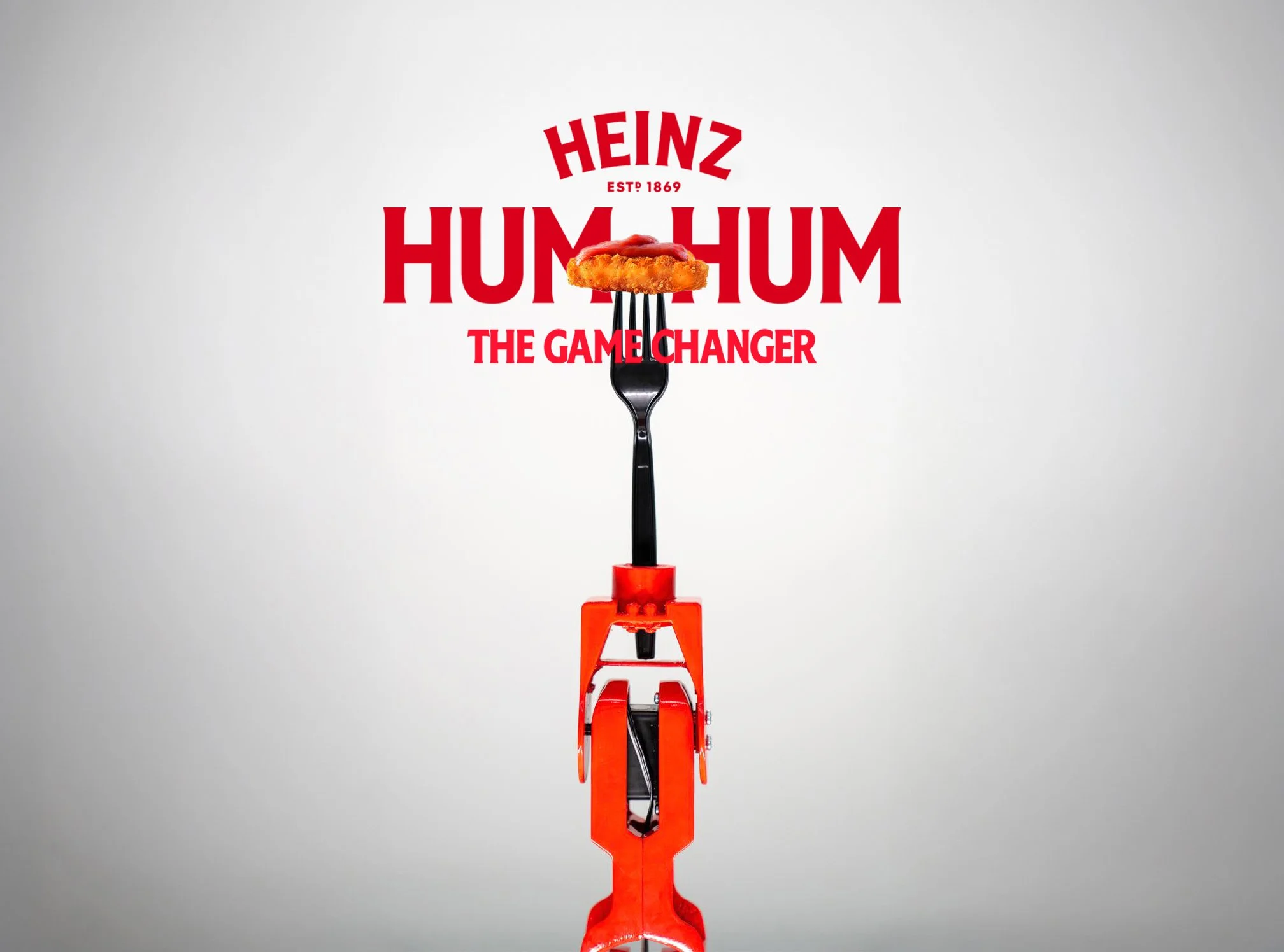 Heinz advert for Hum Hum with a fork balancing a chicken nugget with ketchup on top, centered above an orange toy gun, against a white background.