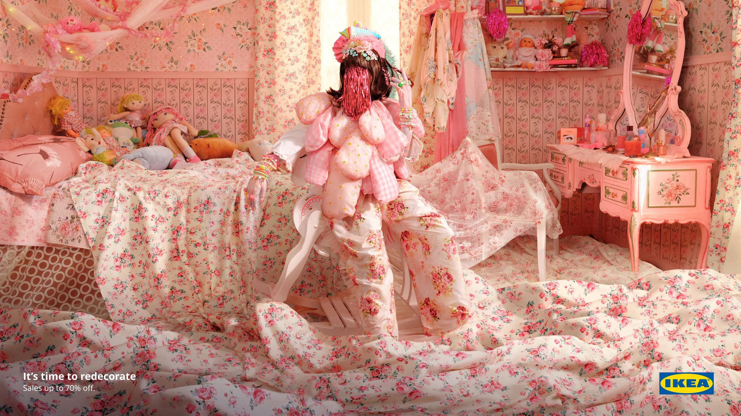 A person dressed in pink and white, wearing a hat and beads, stands in a bedroom with pink floral decor, surrounded by dolls, plush toys, and vintage furniture, with an IKEA logo at the bottom right corner.