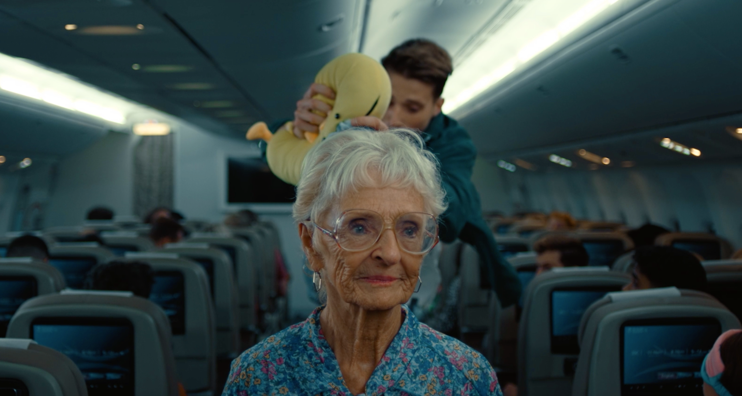 An elderly woman with white hair and glasses sitting on an airplane, with a young man behind her holding a yellow stuffed animal over her head, inside an airplane cabin with seatback screens visible.