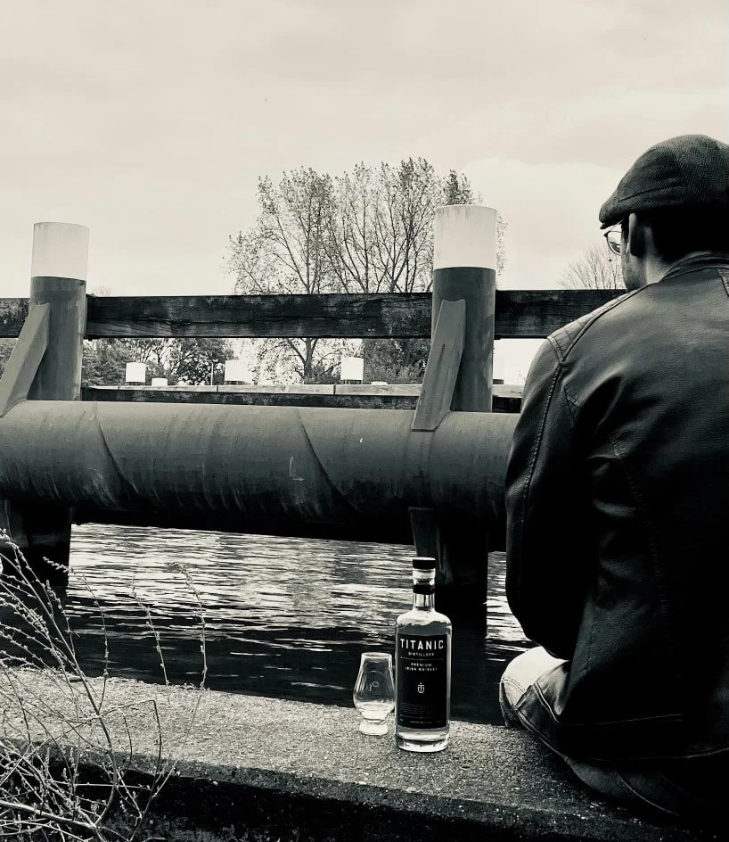 A man in a leather jacket and cap sitting by a canal, looking away, with a bottle of Titanic gin and a glass on the ledge beside him, trees in the background.