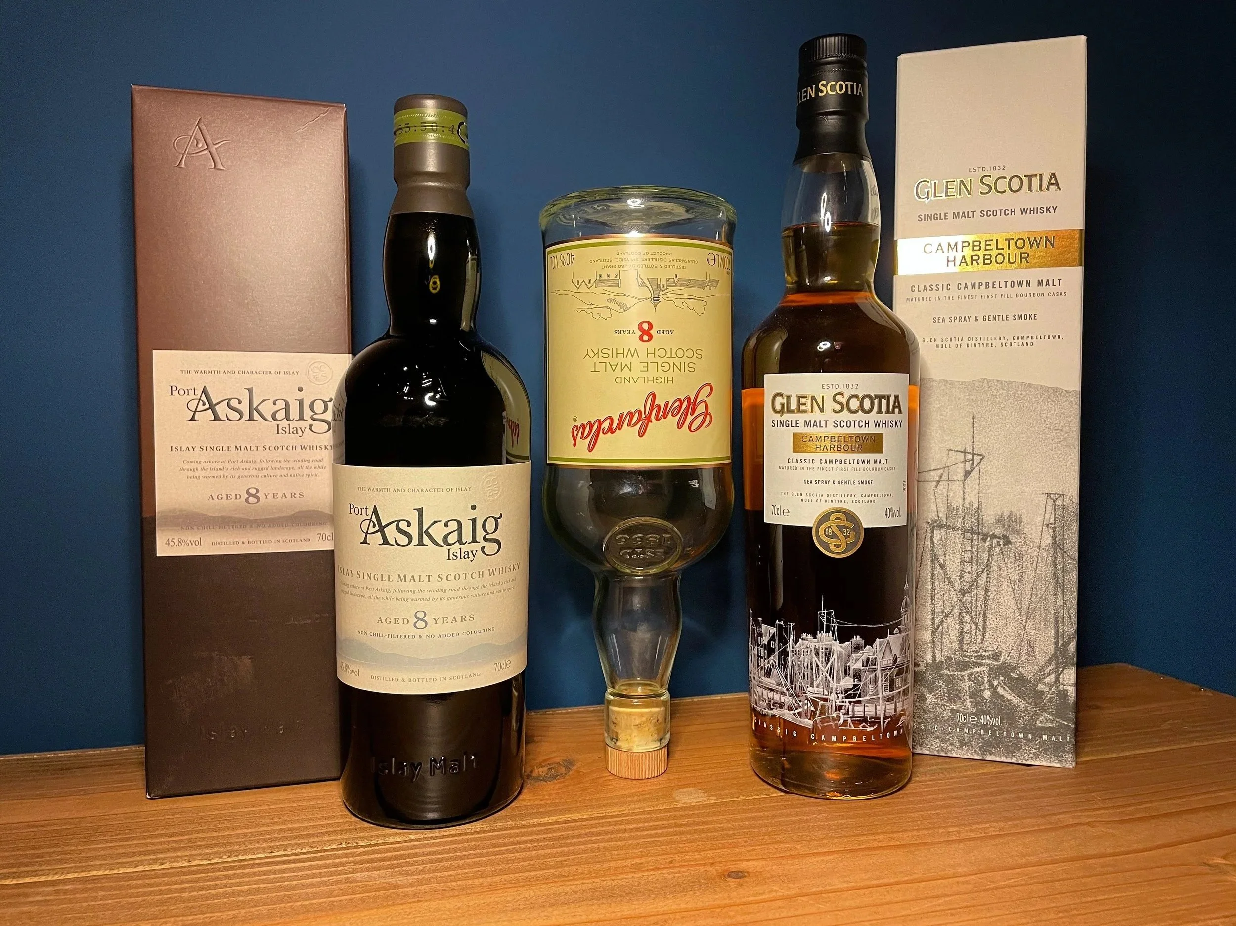 Two bottles of Portaskaig Islay single malt Scotch whisky with black caps and labels, a half-filled glass with whisky, and boxes for each whisky on a wooden surface against a blue wall.