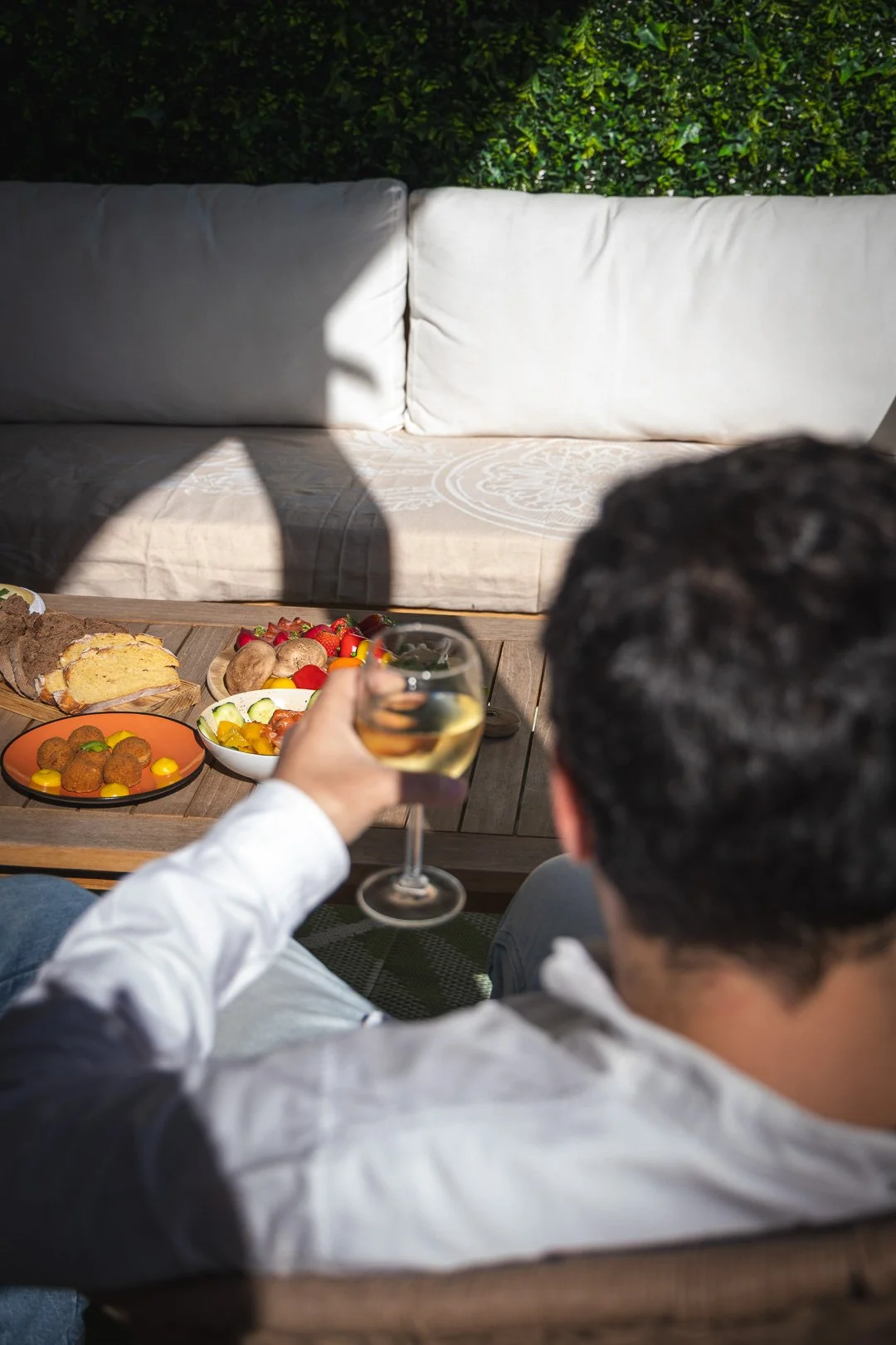 Pessoa segurando taça de vinho branco, com mesa de comida ao fundo, incluindo salada, pão e almôndegas, em ambiente ao ar livre com sofá e plantas