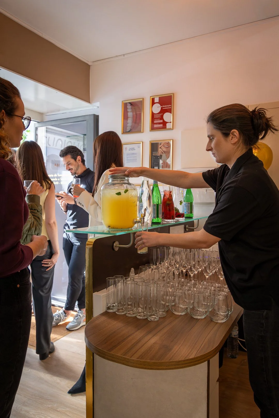 Cozinheira preparando bebidas em balcão, enquanto clientes aguardam dentro de um café ou bar