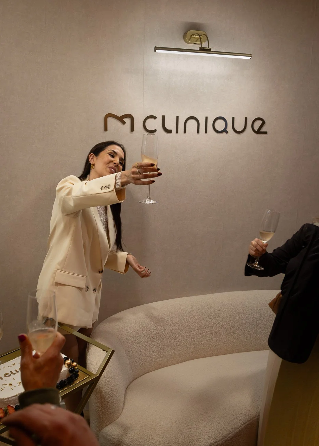 Mulher sorridente com blazer bege segurando uma taça de champagne, ao fundo há uma parede com a placa 'MClineque' e outras pessoas também segurando taças, num ambiente sofisticado.