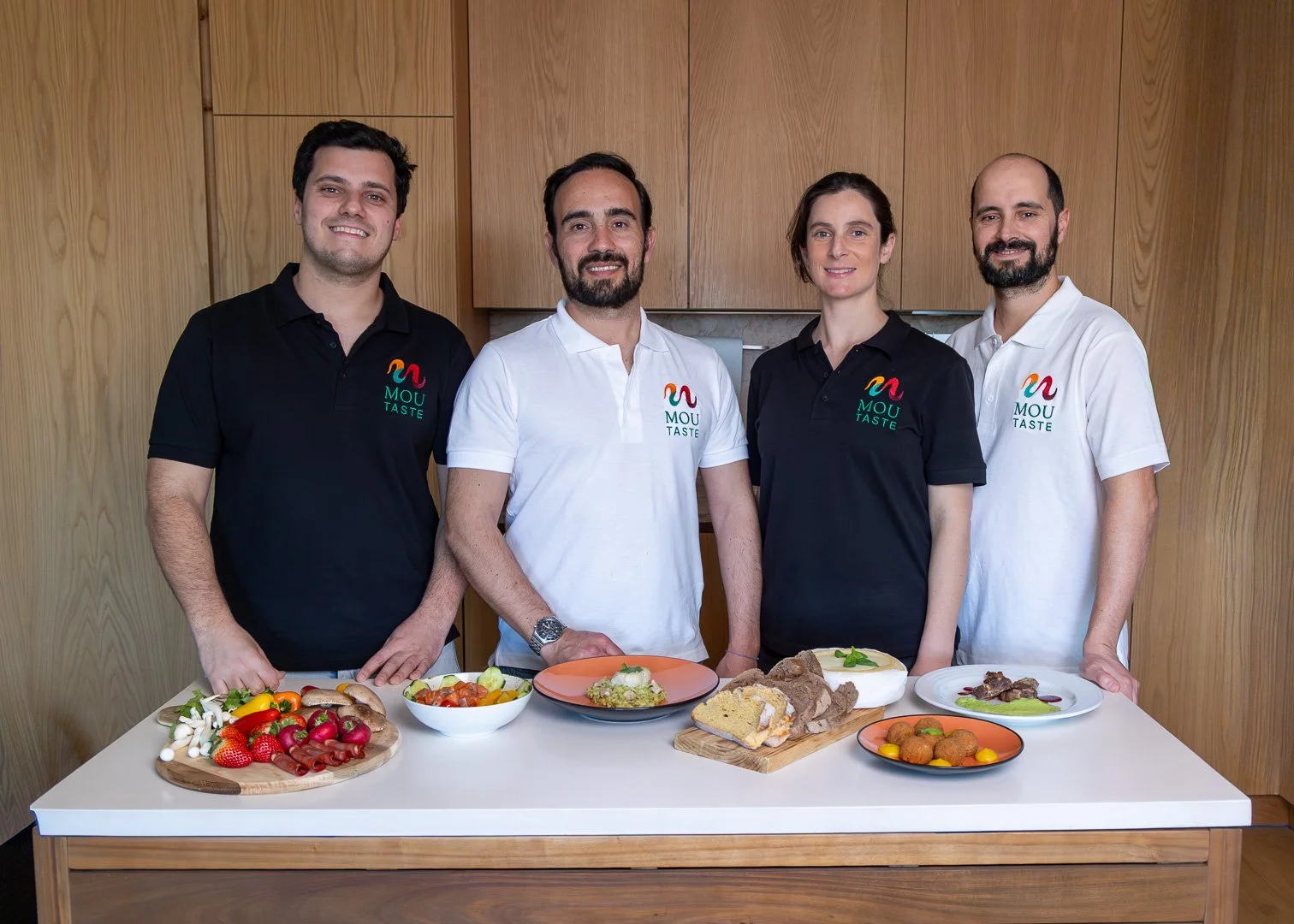 Membros da equipa da Mou Taste. Quatro pessoas de camiseta preta e branca com o logo 'MOU TASTE' posando atrás de uma mesa com pratos de comida