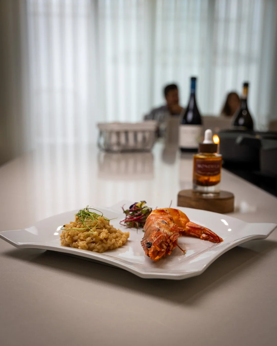 Prato de peixe com arroz e salada, ao fundo garrafas de vinho, um frasco de óleo e pessoas ao fundo em um ambiente interno.