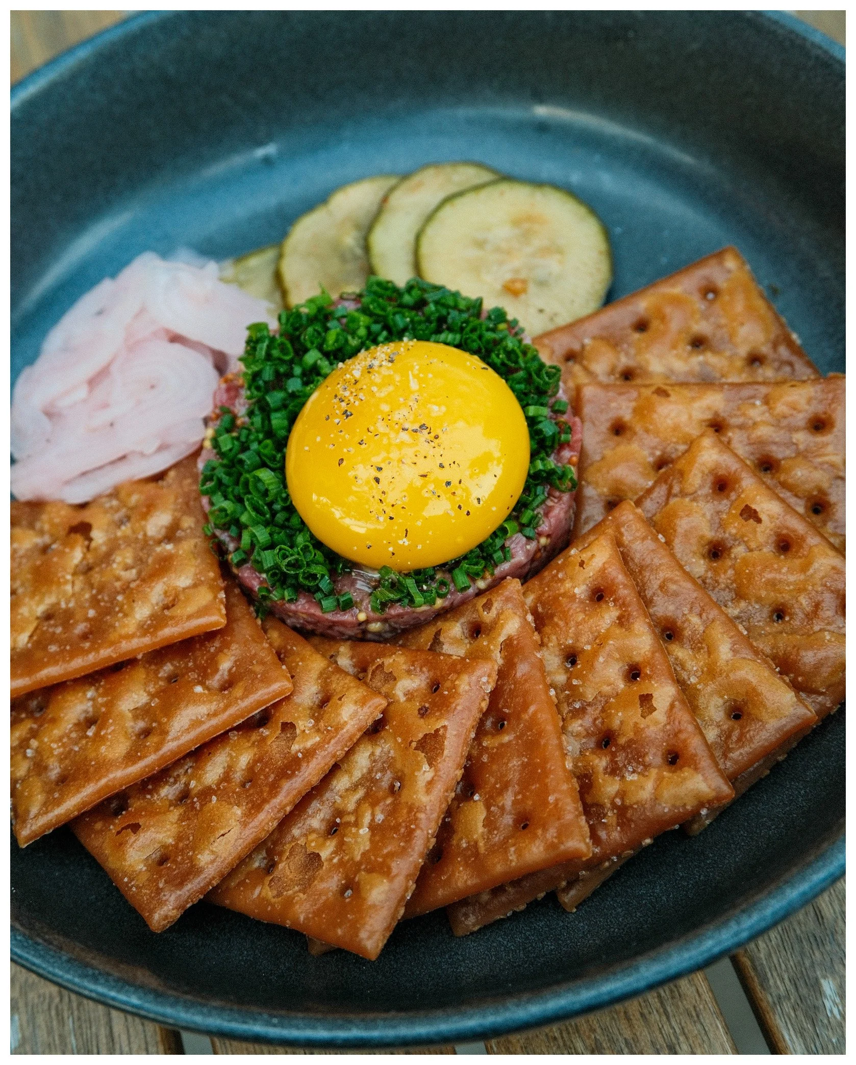 Bite into the perfect beef tartare topped with a raw egg yolk, surrounded by crispy crackers, sliced cucumbers, pickled onions, and chopped green onions, served on a dark blue plate.