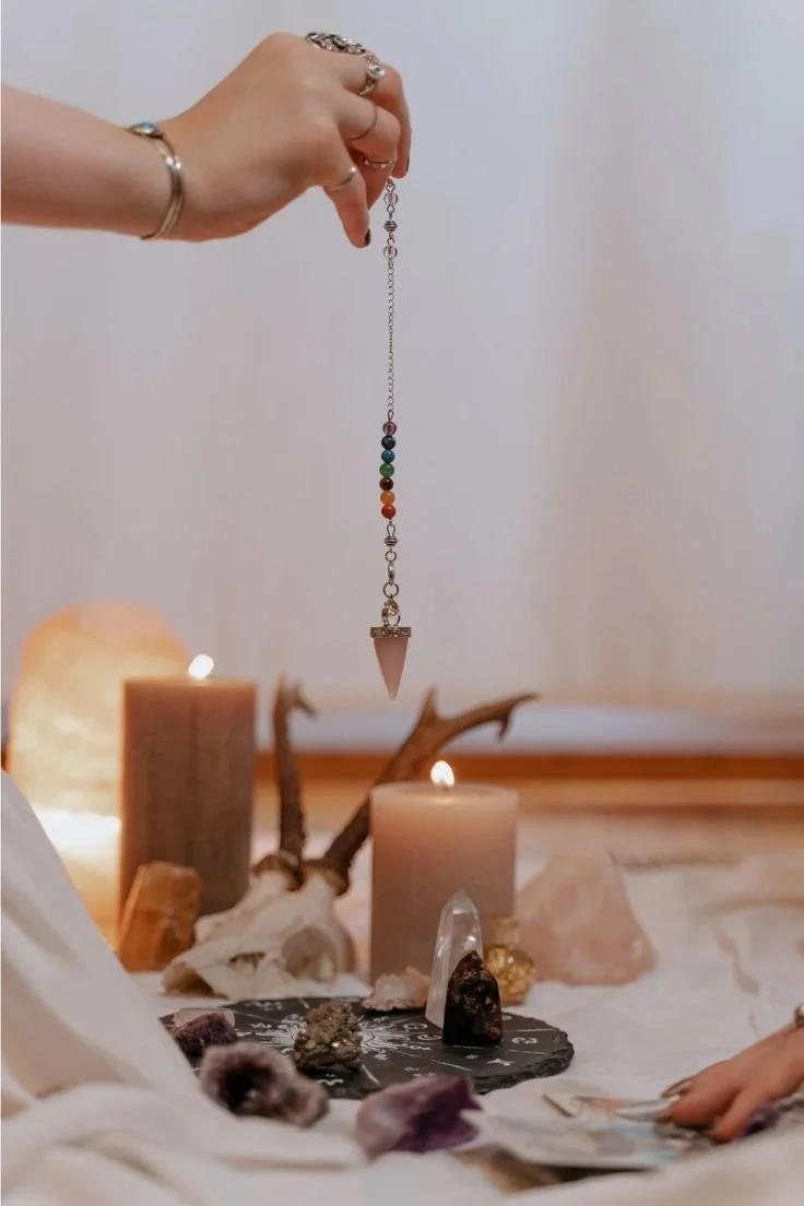 Une main tient un pendule de cristal avec un symbole d'arc-en-ciel de sept couleurs en pendentif, suspendu au-dessus d'une surface avec des bougies, des cristaux et des objets de décoration. L'ambiance est calme et mystérieuse.