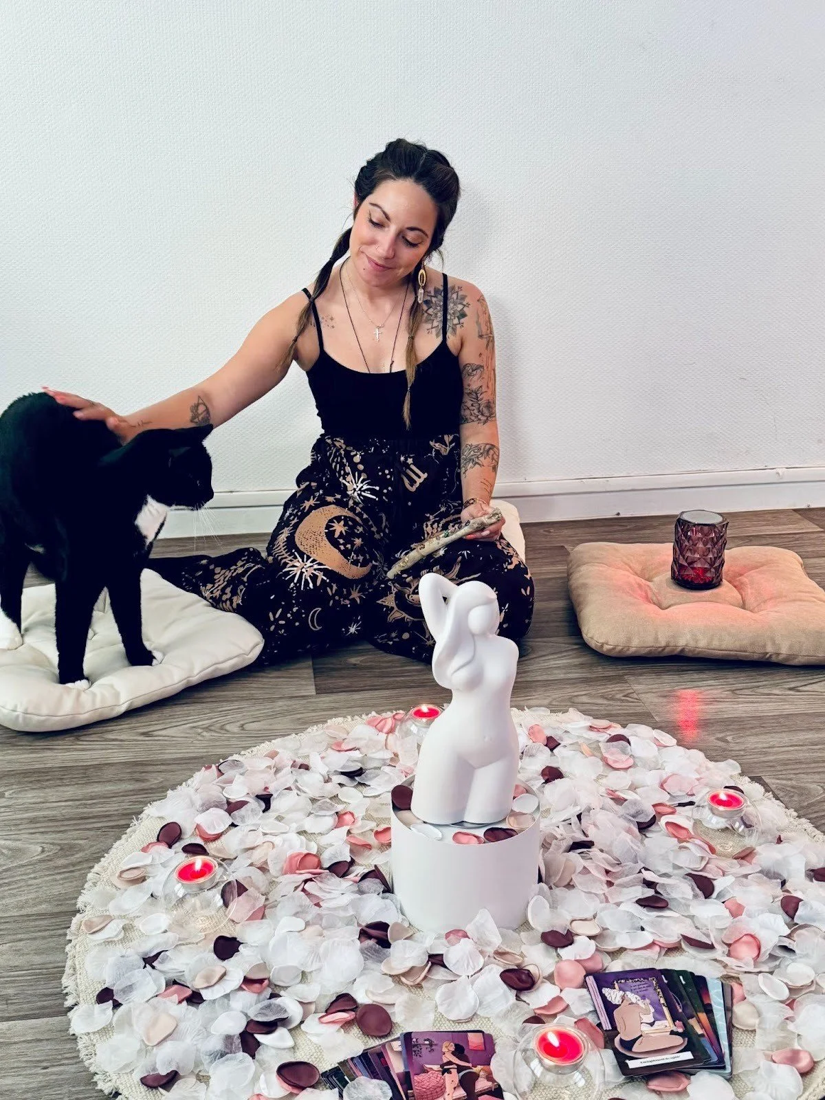 Une femme assise sur le sol, entourée de pétales, avec une chatte noire et blanche entre ses jambes. Une statue de femme et plusieurs bougies sont présentes, avec des coussins et un verre violet à l'arrière.
