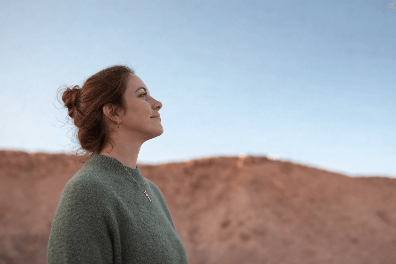 Femme avec des cheveux bruns en chignon, portant un pull vert foncé et une croix en collier, regarde vers le ciel dans un paysage désertique au coucher du soleil.
