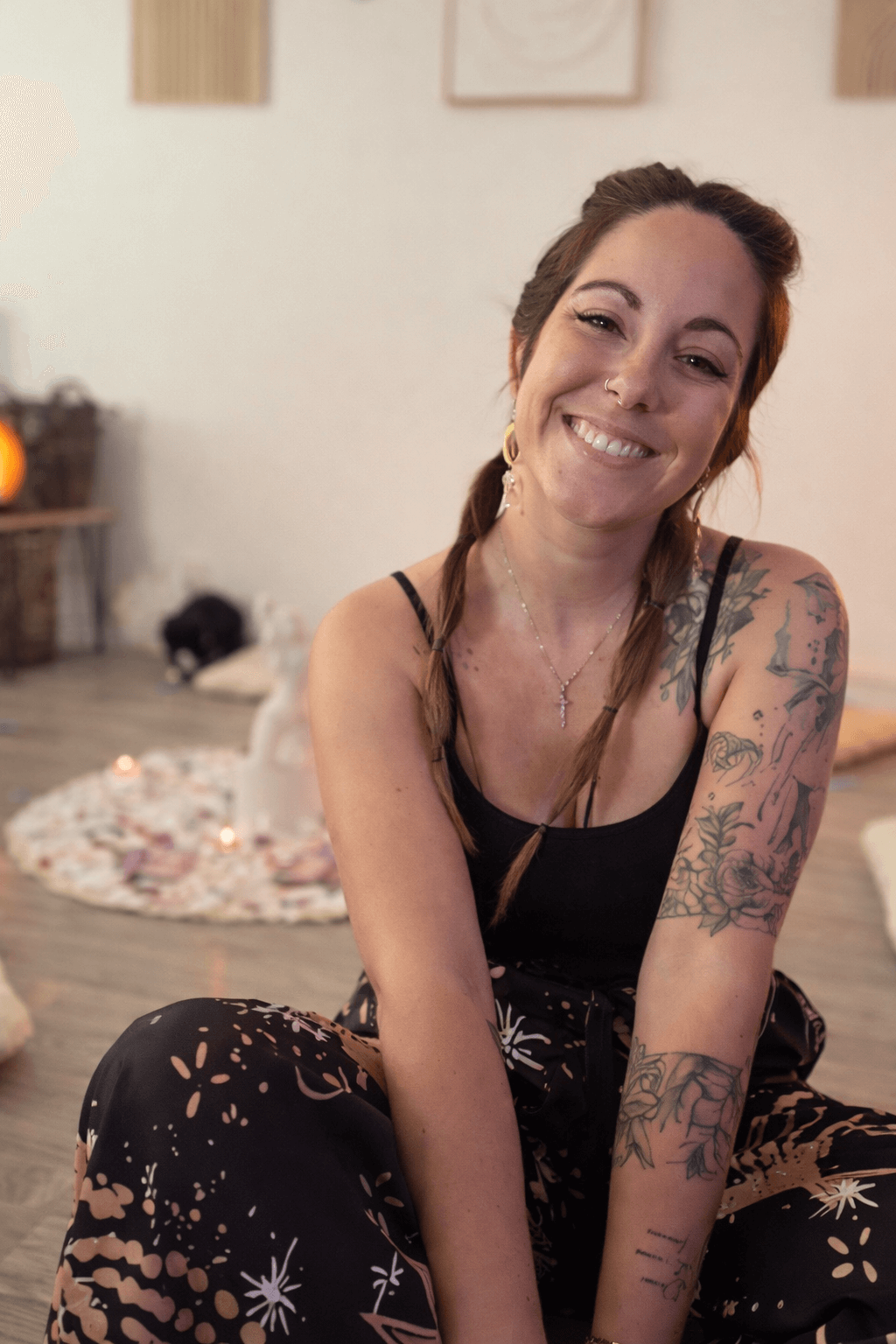 Jeune femme souriante avec des tatouages, portant un haut noir, assise à l'intérieur, avec un décor chaleureux et des bougies en arrière-plan.