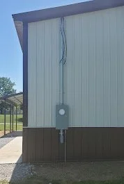 Exterior corner of a building with a light-colored wall, dark base trim, and an electrical box mounted on the wall, with some wires hanging down.