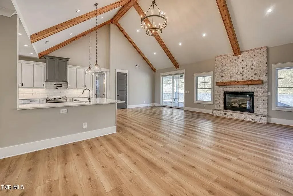 Open living room and kitchen area with vaulted ceiling, wooden beams, and hardwood floors. Fireplace with brick surround and wooden mantel. Sliding glass door and windows allow natural light.