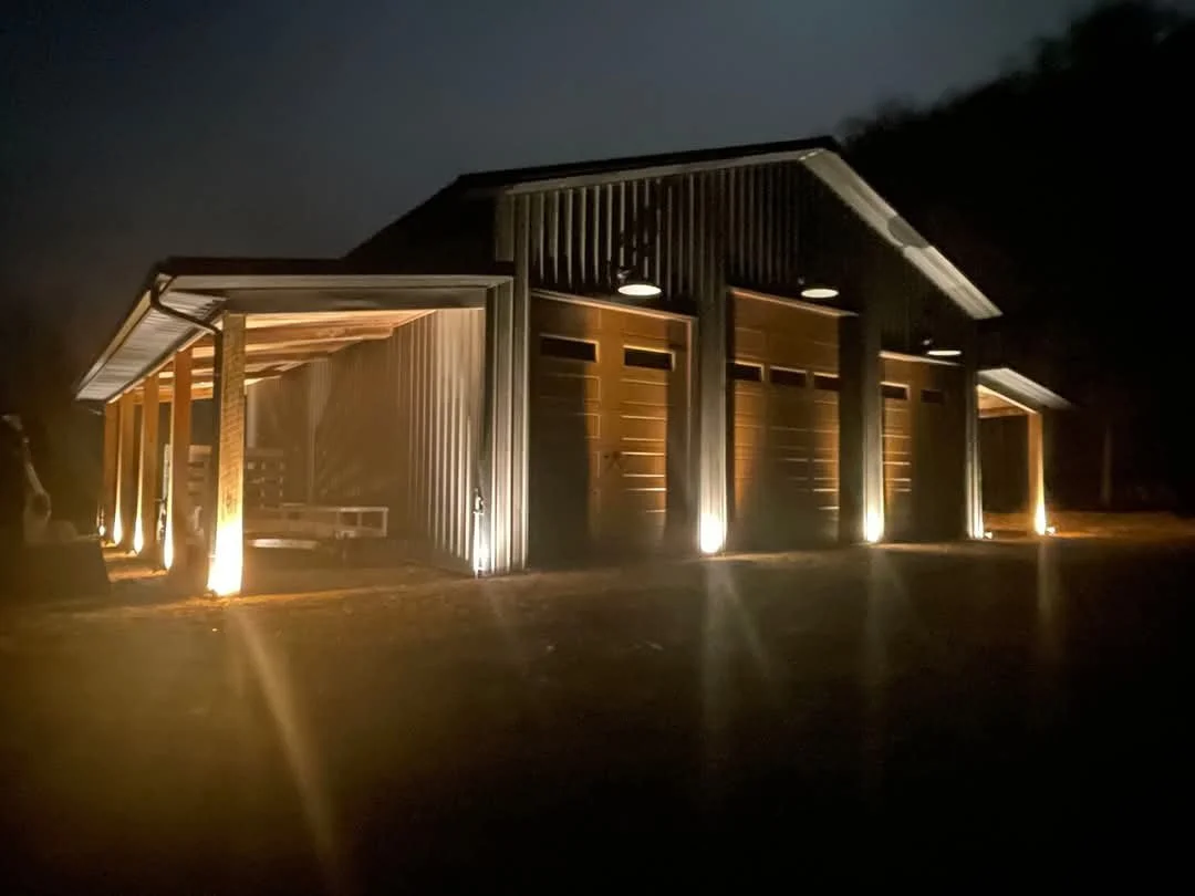 A modern barn-style building with exterior lighting at night, featuring vertical metal siding and large garage doors.