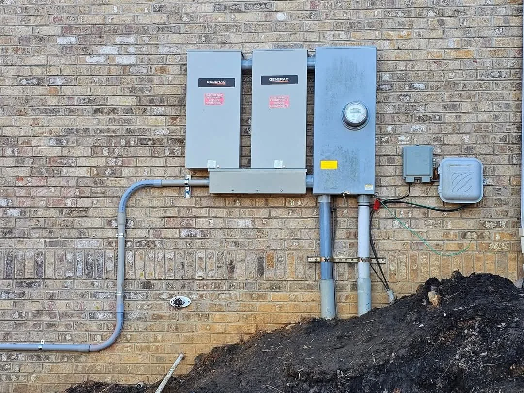 Electrical meter and upgrade boxes mounted on a brick wall, with conduit pipes and a patch of dirt below.