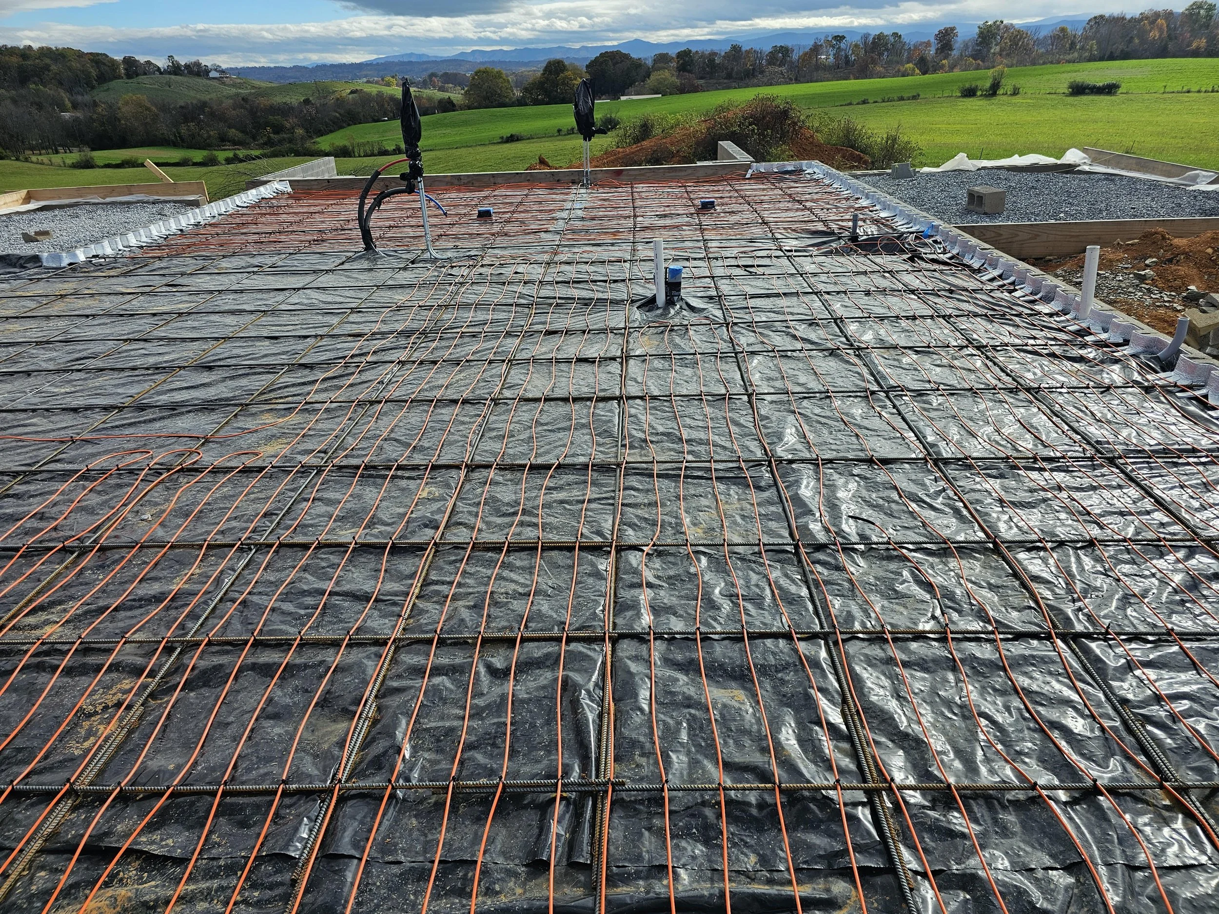 Construction site with electrical wiring and plumbing pipes on a building's roof, overlooking green fields and hills in the background.