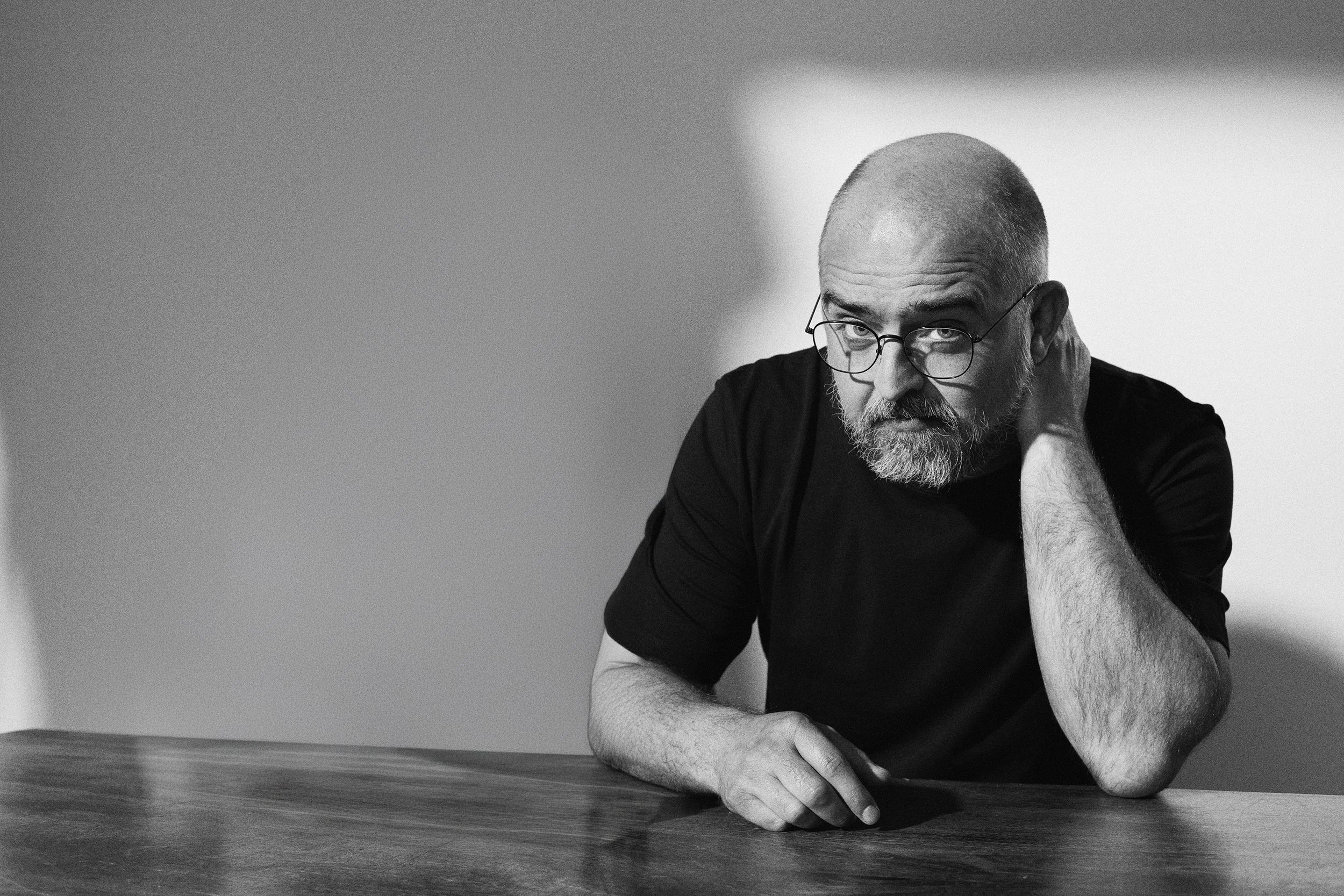 Zittende man met een kale kop, baard, bril en een zwart shirt, rustend zijn hoofd op zijn hand, op een houten tafel, in zwart-wit.