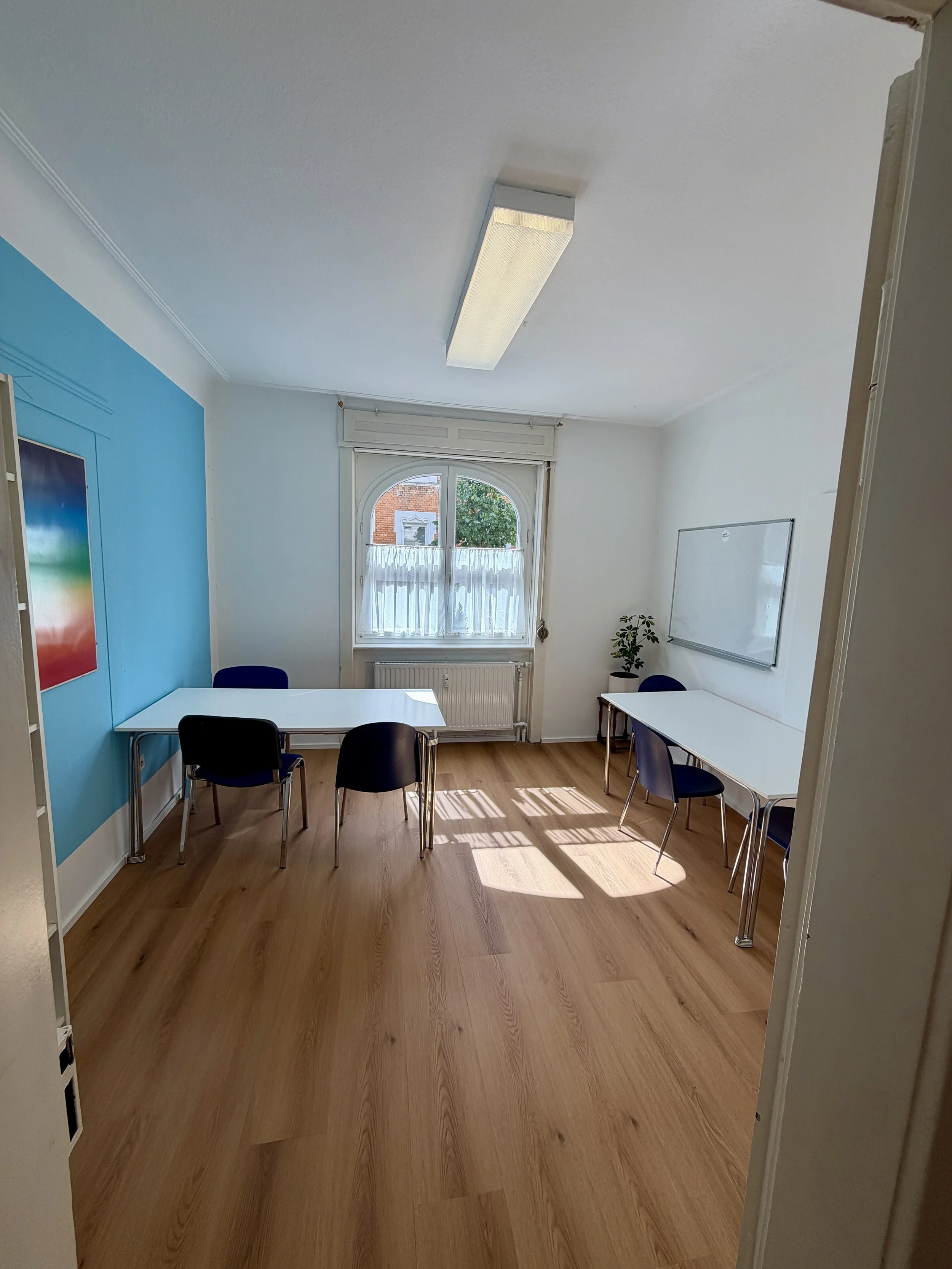 Ein heller, leerer Besprechungsraum mit weißen Wänden, Holzboden, einem großen Fenster mit weißen Vorhängen, einem Whiteboard an der Wand, einem Tisch mit sieben Stühlen und einer Pflanze in der Ecke.