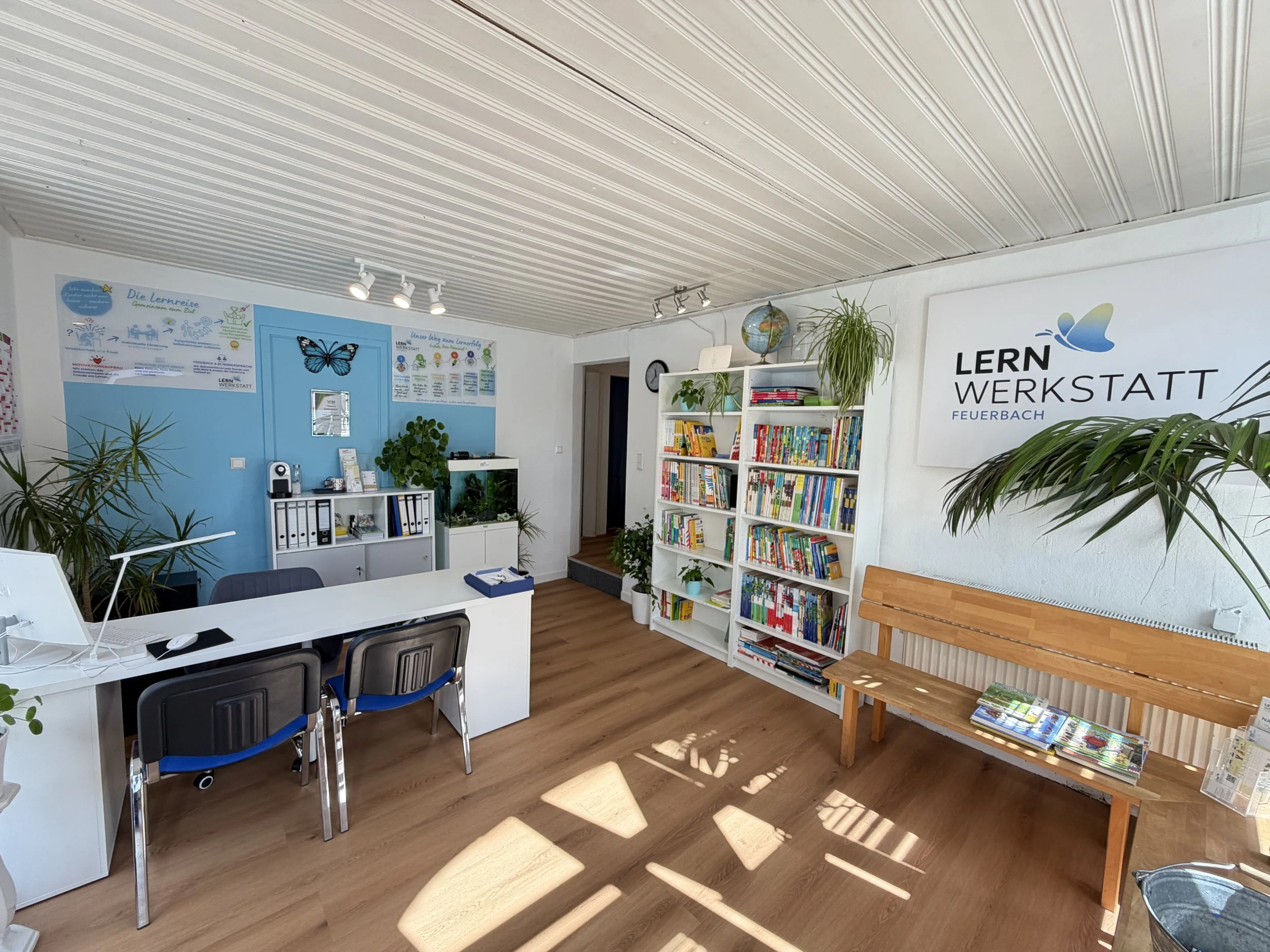 Büro oder Lernraum mit Bücherregalen, Pflanzen, einem Tisch mit Stühlen, einem Plakat an der Wand, und einer Decke mit Holzbalken. Markenname "LERN WERKSTATT Feuerbach" an der Wand.