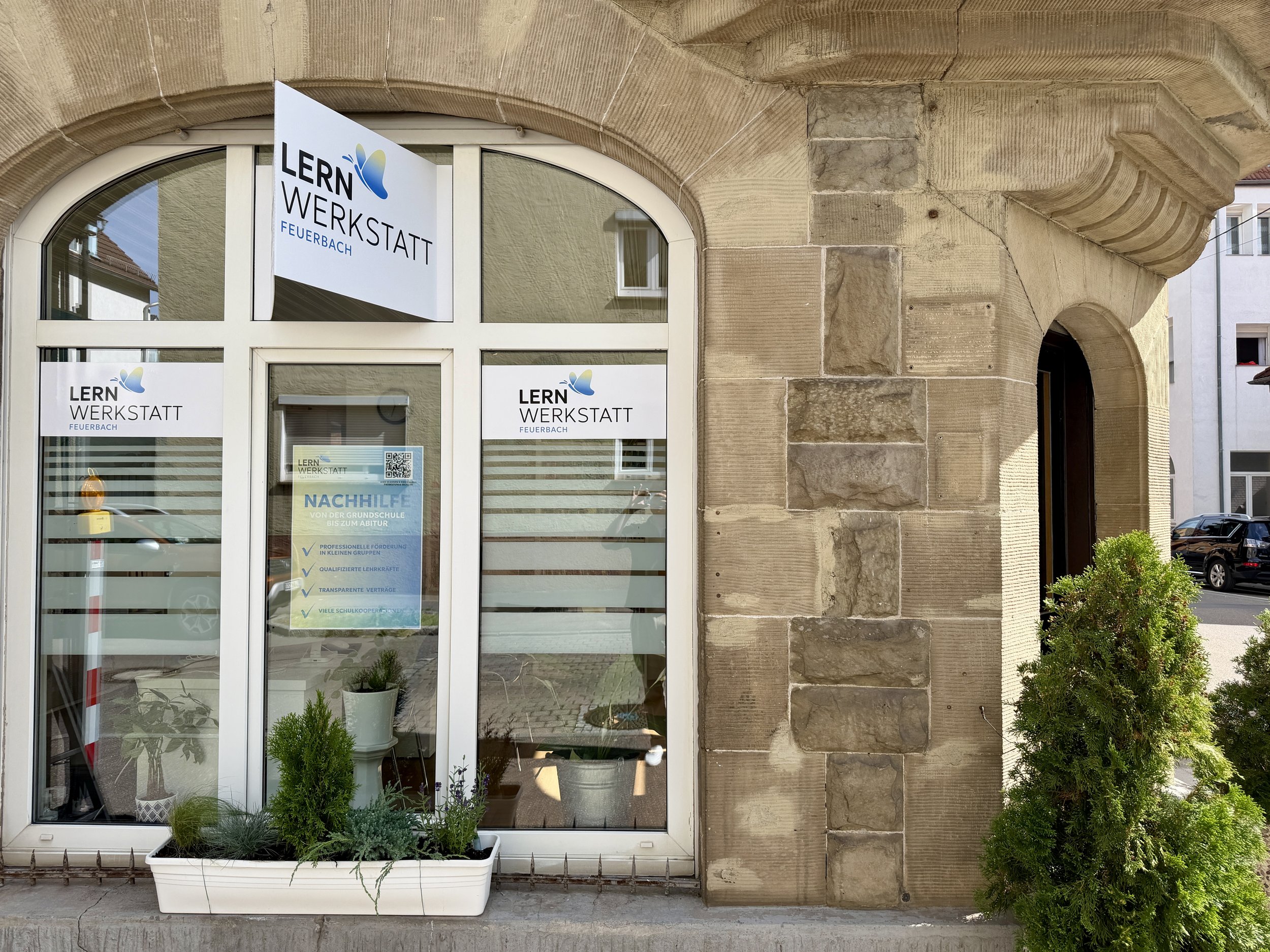 Fenster eines Gebäudes mit Schildern, die 'LERN WERKSTATT FEUERBACH' und ein QR-Code anzeigen. Im Fenster steht ein Pflanzentopf mit grünen Pflanzen. Das Gebäude besteht aus einem historischen Steinmauerabschnitt.