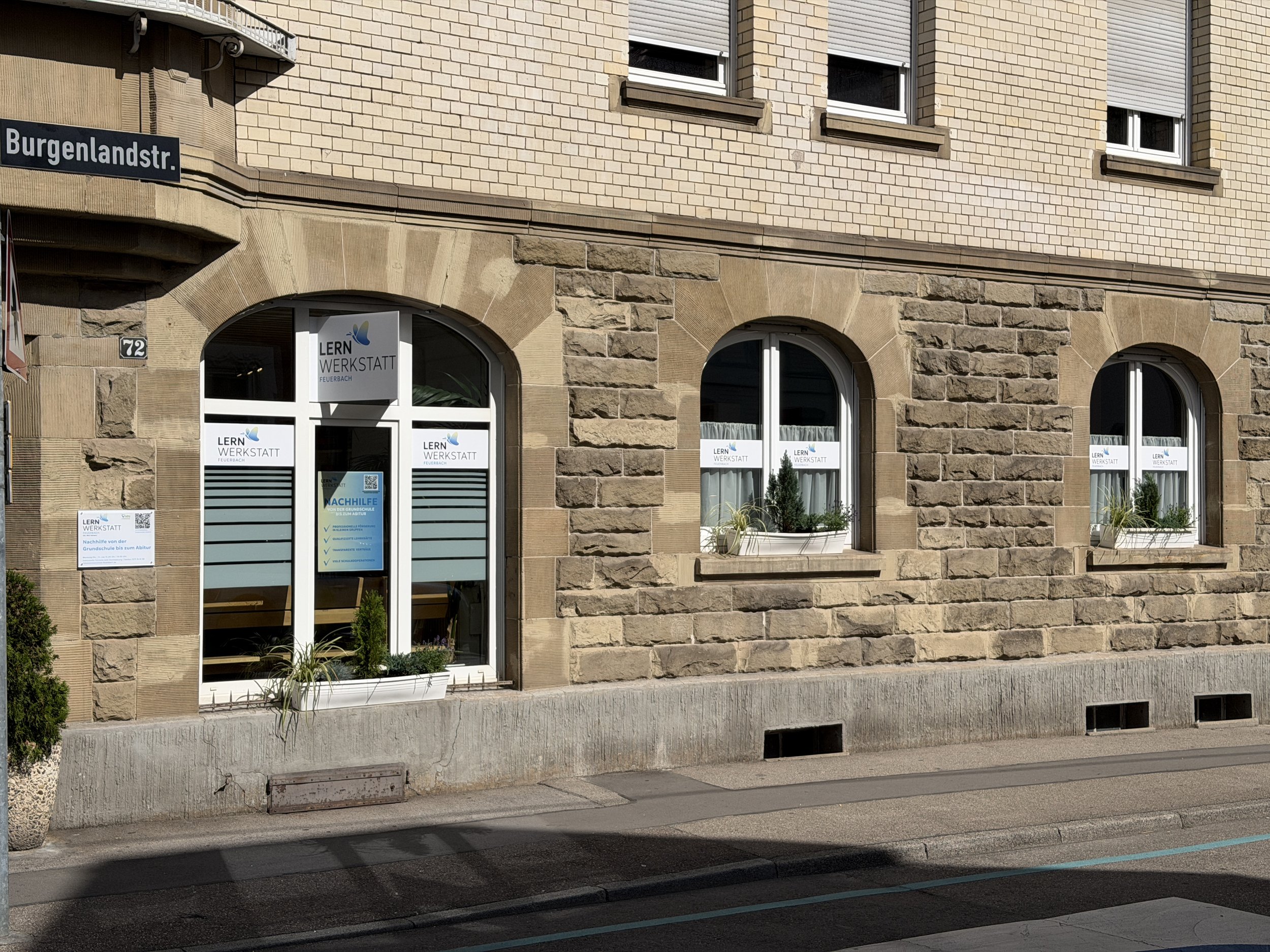 Das Bild zeigt die Fassade eines Gebäudes mit drei Stockwerken. Es hat Fenster mit weißen Rahmen und Shutters, einige davon sind geschlossen. Das Erdgeschoss hat teilweise geschwungene Tür- und Fensterrahmen aus weißem Material, mit Pflanzen in Pottkübeln davor. Es gibt ein Straßenschild mit der Aufschrift 'Burgenlandstr.' und eine weitere Tafel mit dem Logo 'LERN WERKSTATT Feuerbach' im unteren Bereich der Tür.