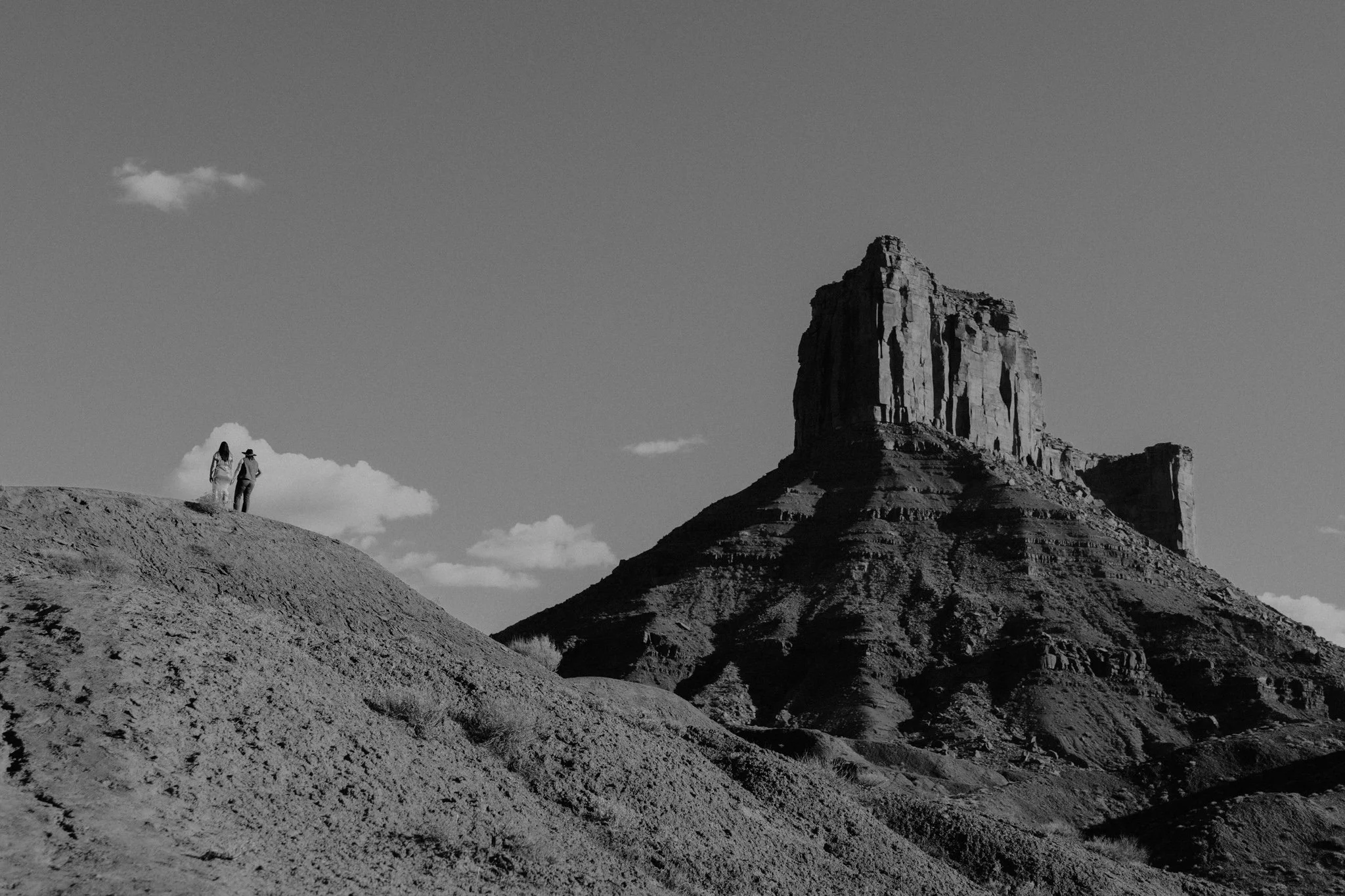 Black and white photo of a queer couple, far away, holding hands with a large Moab mesa in the background
