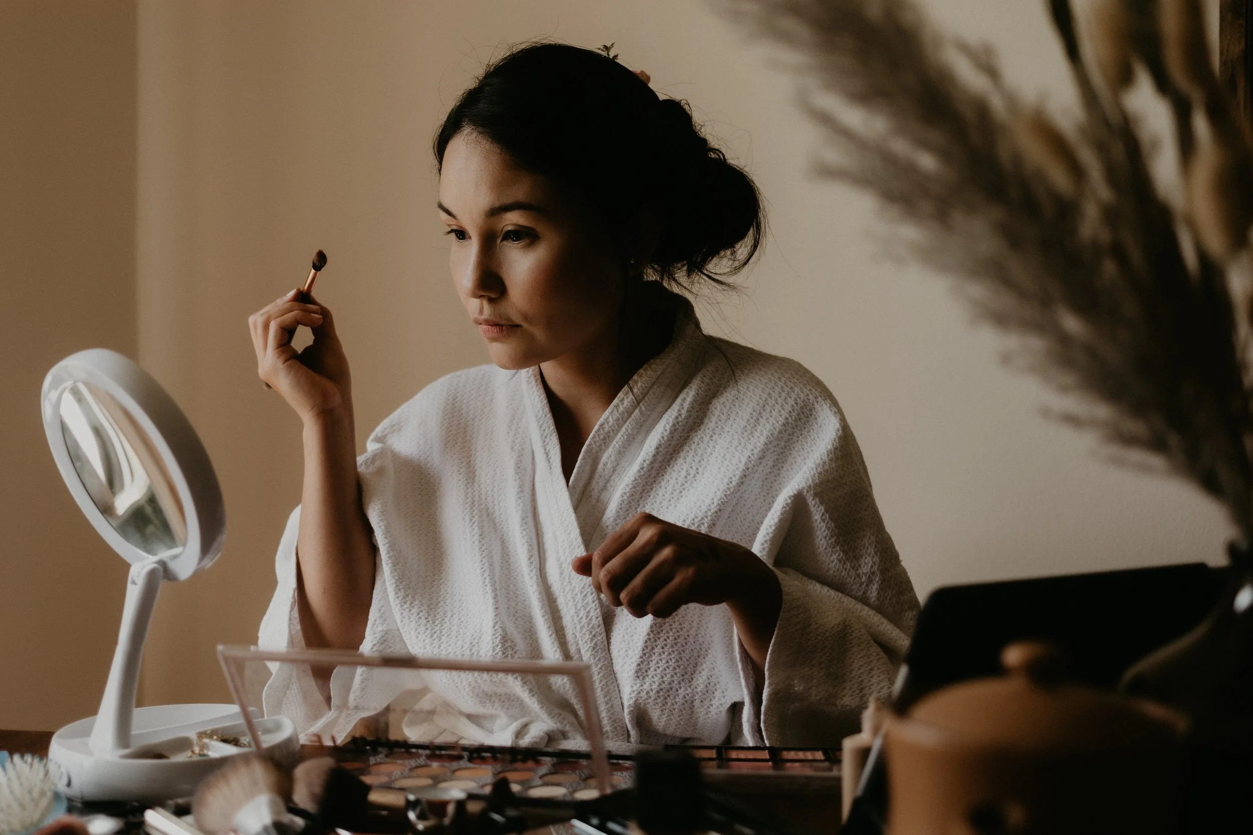 Bride looks into a small mirror, makeup on her table and a small brush in her hand