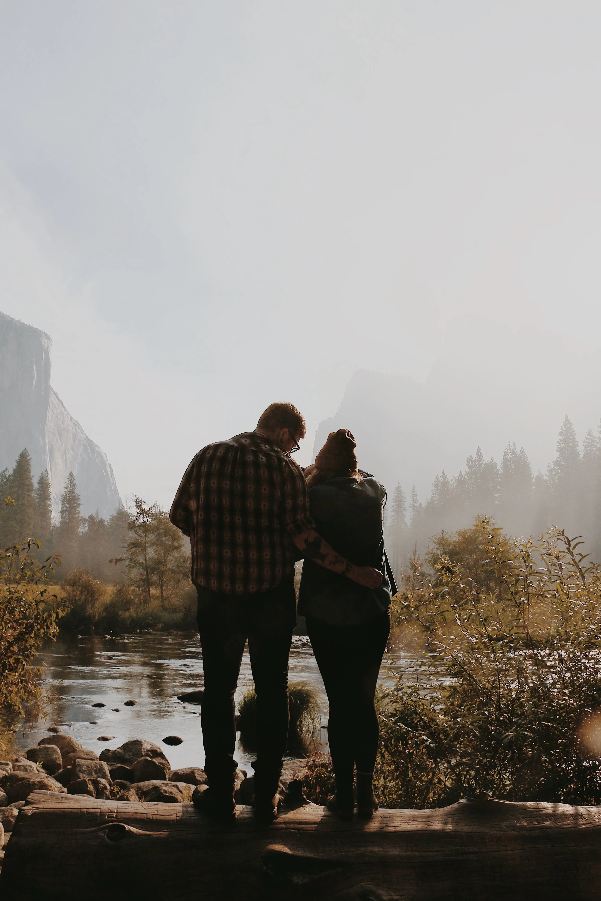 Paige and Corey of Going Home Co. standing on a log at a Yosemite River, embracing and looking at the scenic background of El Capitan, trees and mountains.