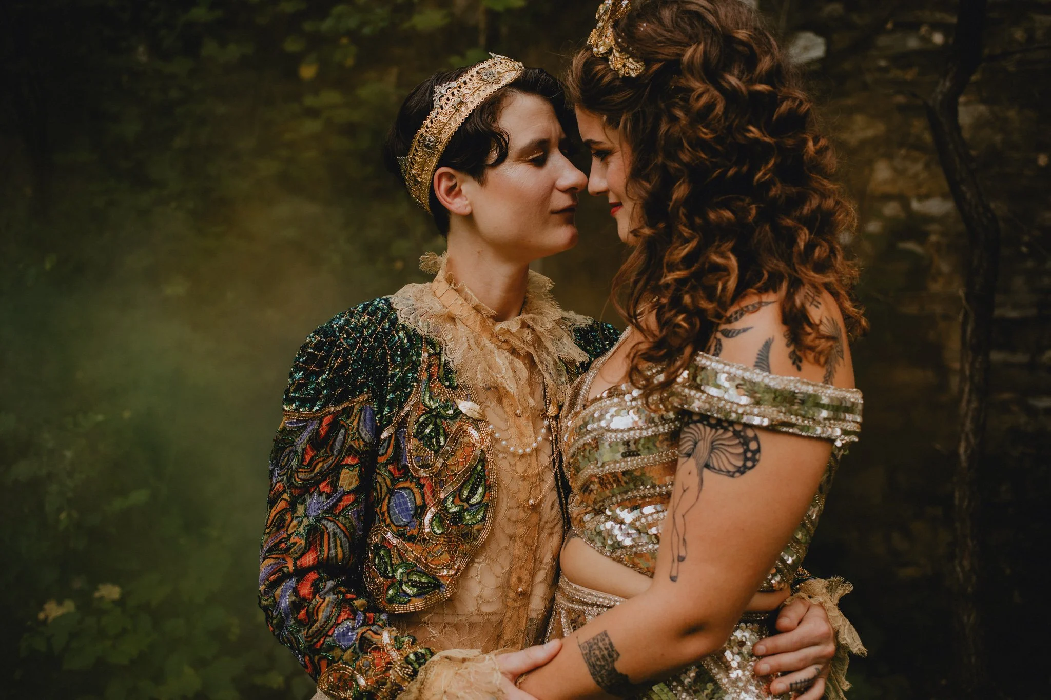 Two brides are nose to nose as they go in for a kiss, in their gold thrifted outfits. A wall of ivy and orange and green smoke bomb behind them.