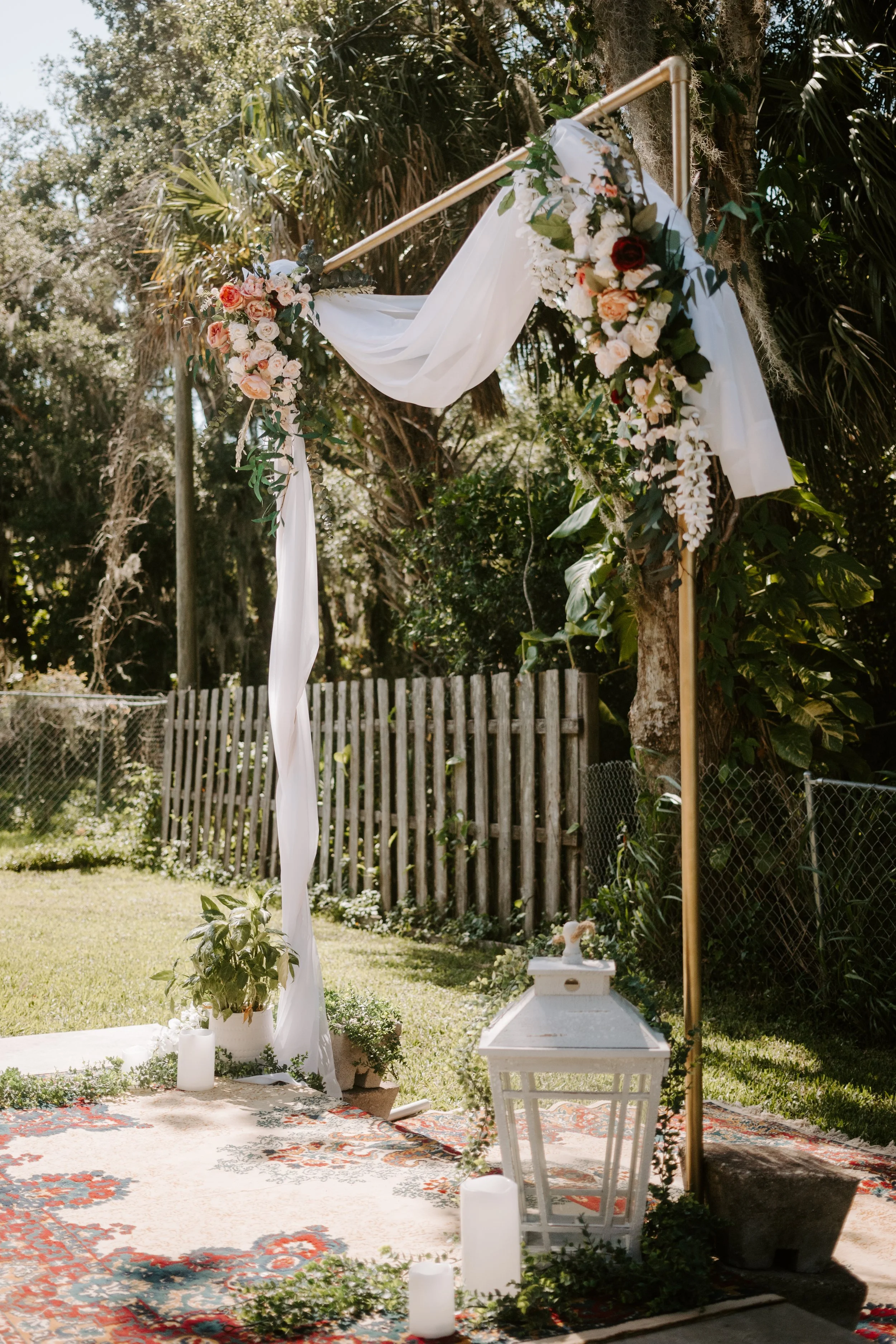 A backyard adorned with palm trees and a wooden fence. A large ornate rug is placed on the ground with a golden arch and diy floral arrangements. Lanterns, live plans and candles are arranged along the base of the arch.