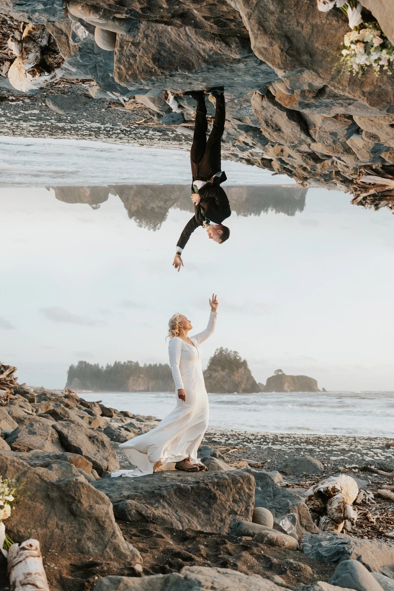 A bride in a white wedding dress standing on a rocky beach, reaching up with her hand; a man in a black suit standing on a rock upside down, overhead, reaching down towards her.
