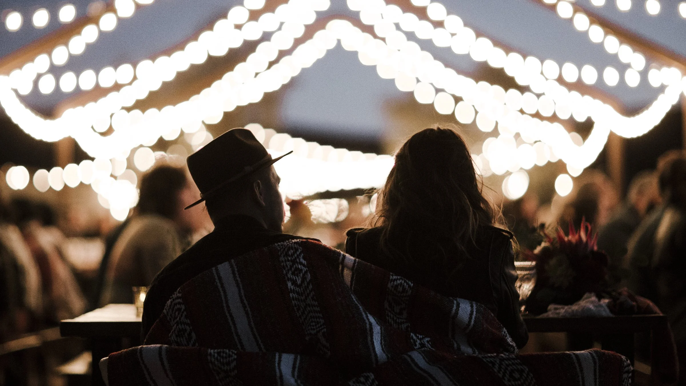 wedding couple, wrapped in a blanket, sits under the exposed beams at the ruin venue with cafe lights above