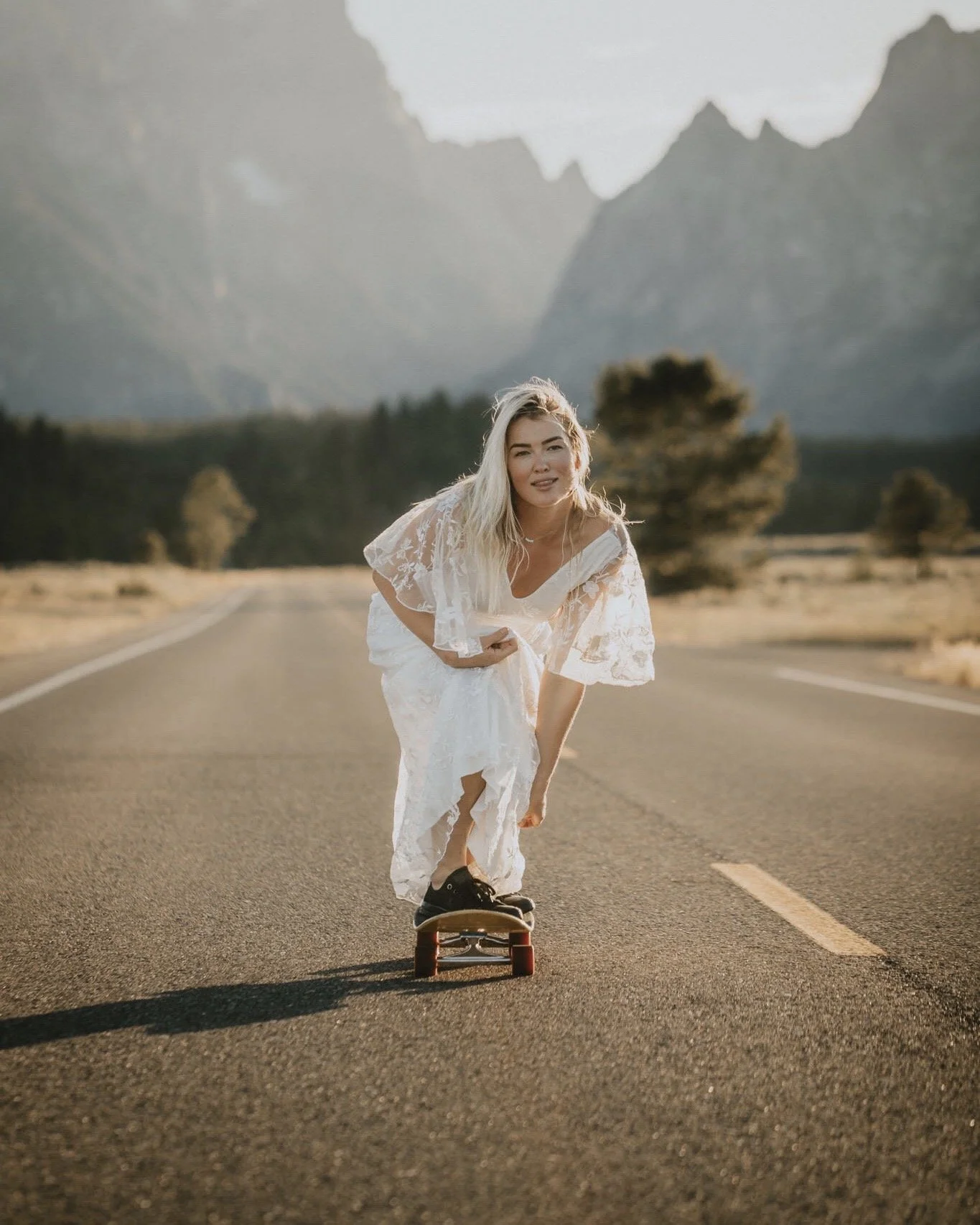 Bride in her wedding dress skateboarding on an empty road with mountains in the background.