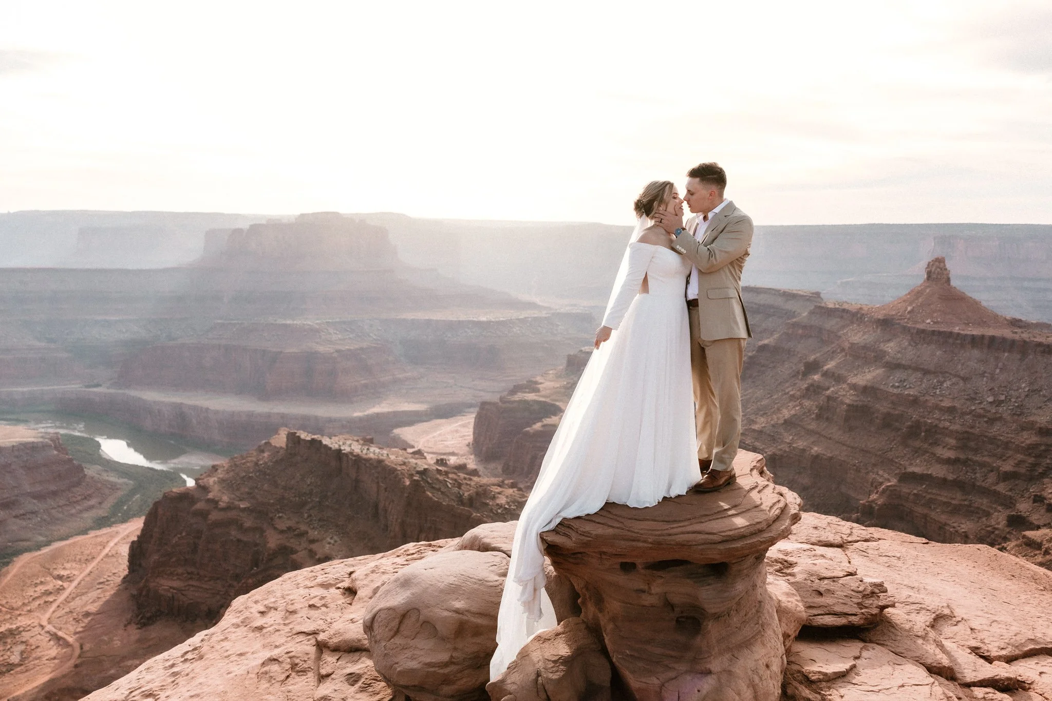 Wedding couple share a kiss standing on a rocky ledge in Dead Horse Point State Park with the setting sun