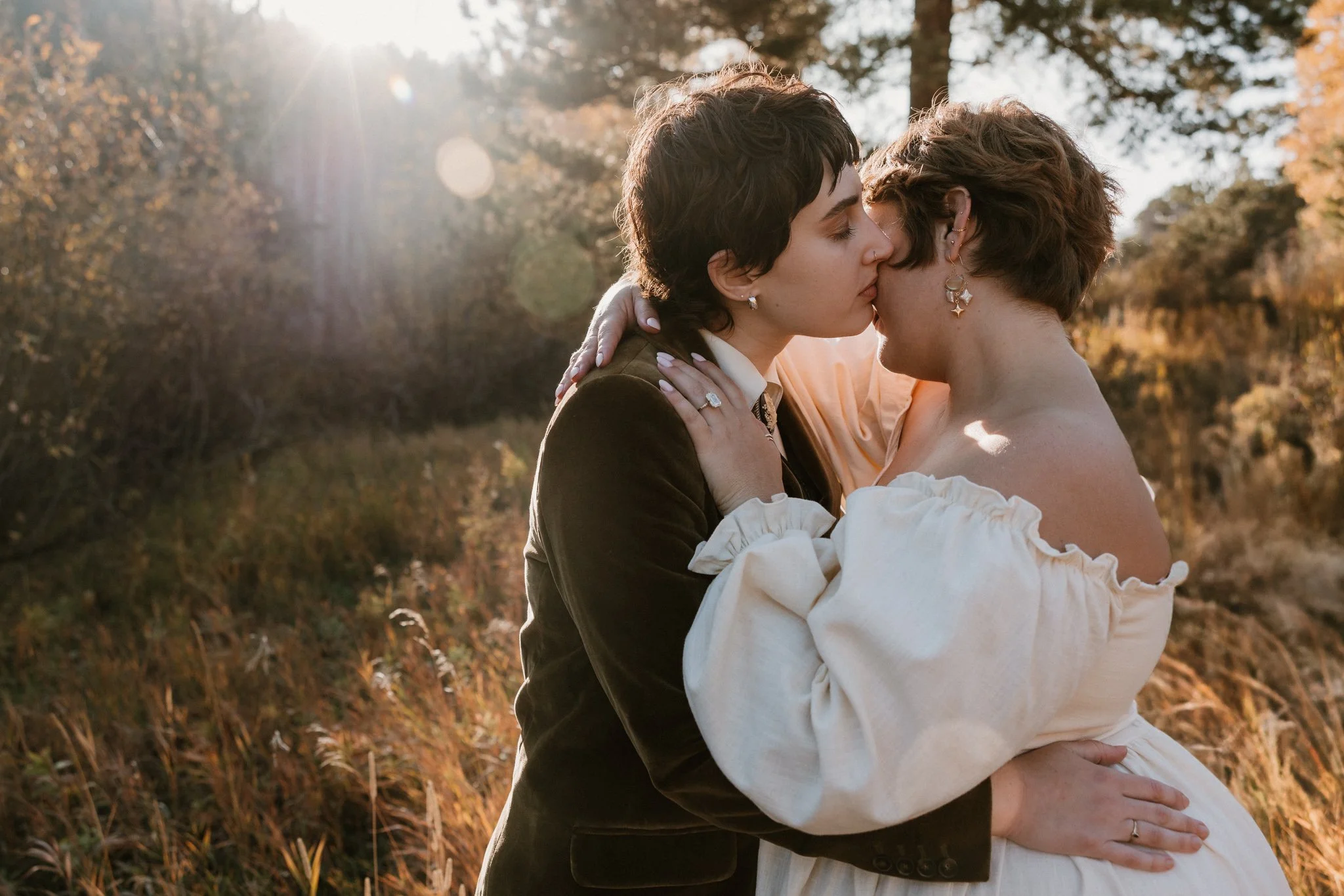Two women kissing outdoors in a field during sunset, with trees and golden light in the background.