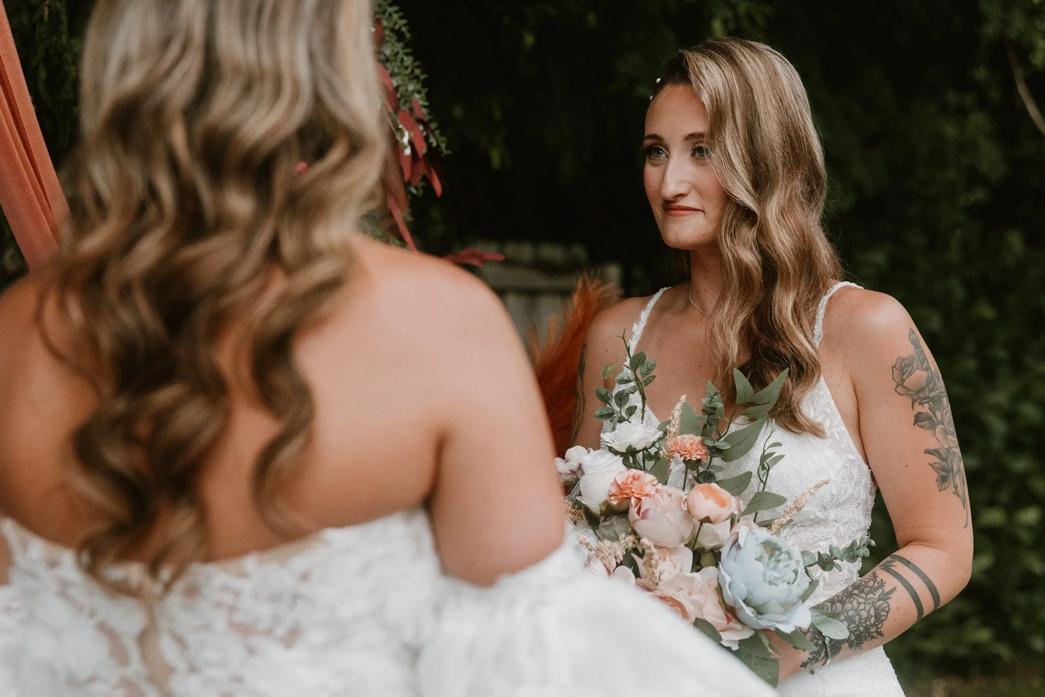 Queer couple at their backyard Florida elopement ceremony. A bride holding a bouquet of white and pink flowers and green foliage, facing another person with shoulder-length wavy hair, outdoors with green foliage background.