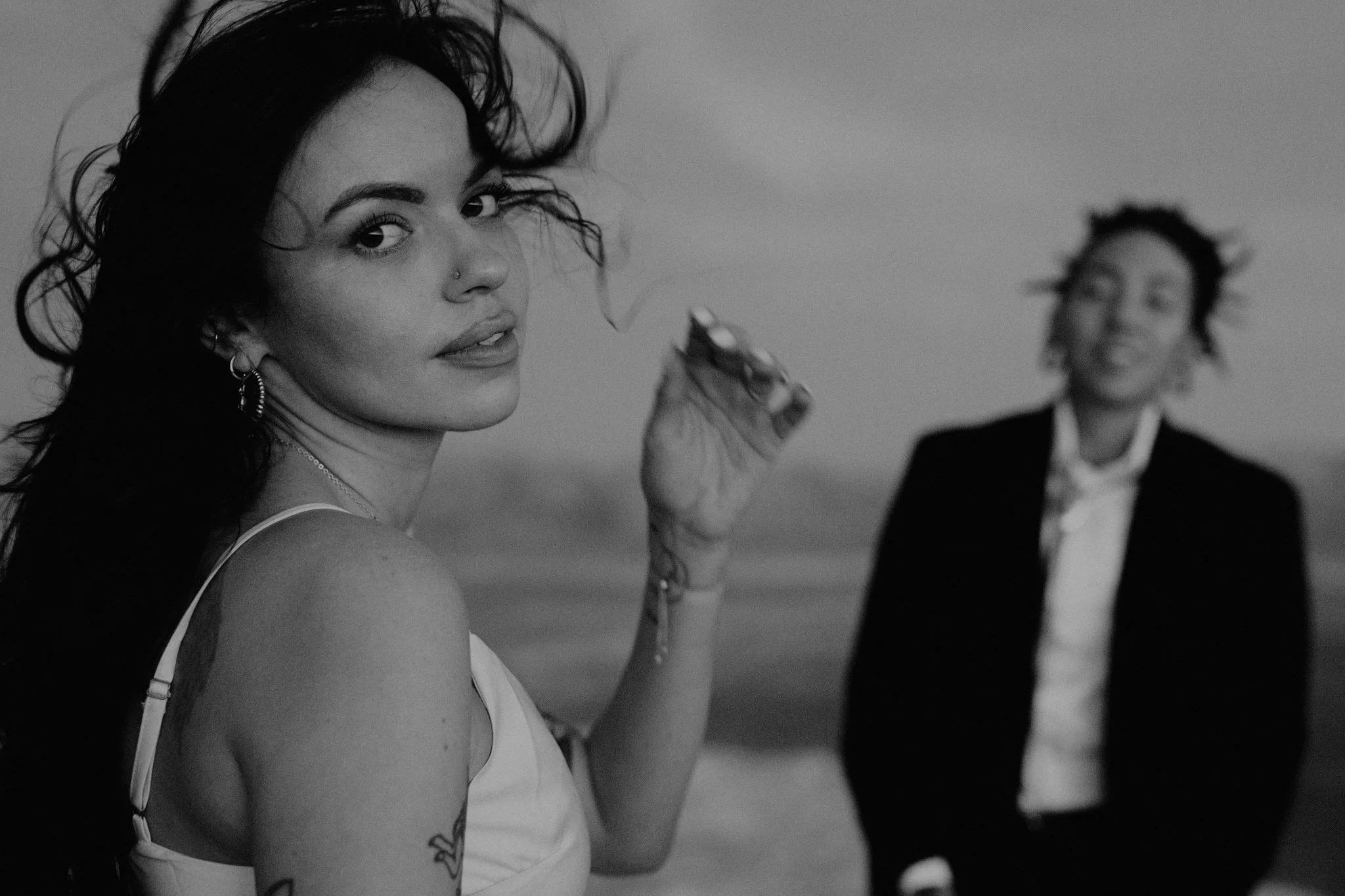 A black and white photo of a queer elopement couple, one bride with dark hair, wearing earrings and a tattoo on her arm, looking at the camera, with a smiling bride in the background.