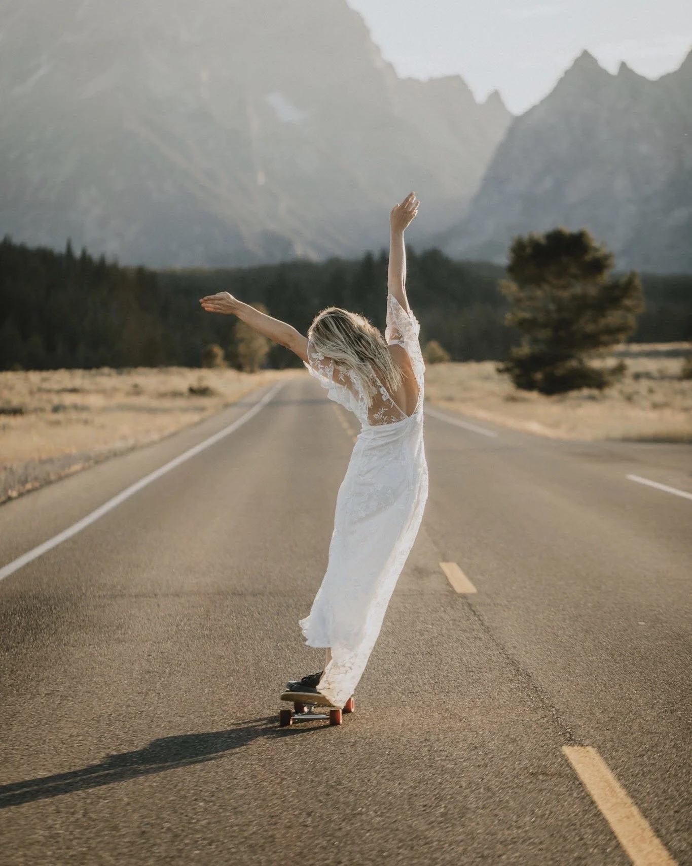 A bride in a white wedding dress skateboarding on an empty road in Grand Tetons National Park.