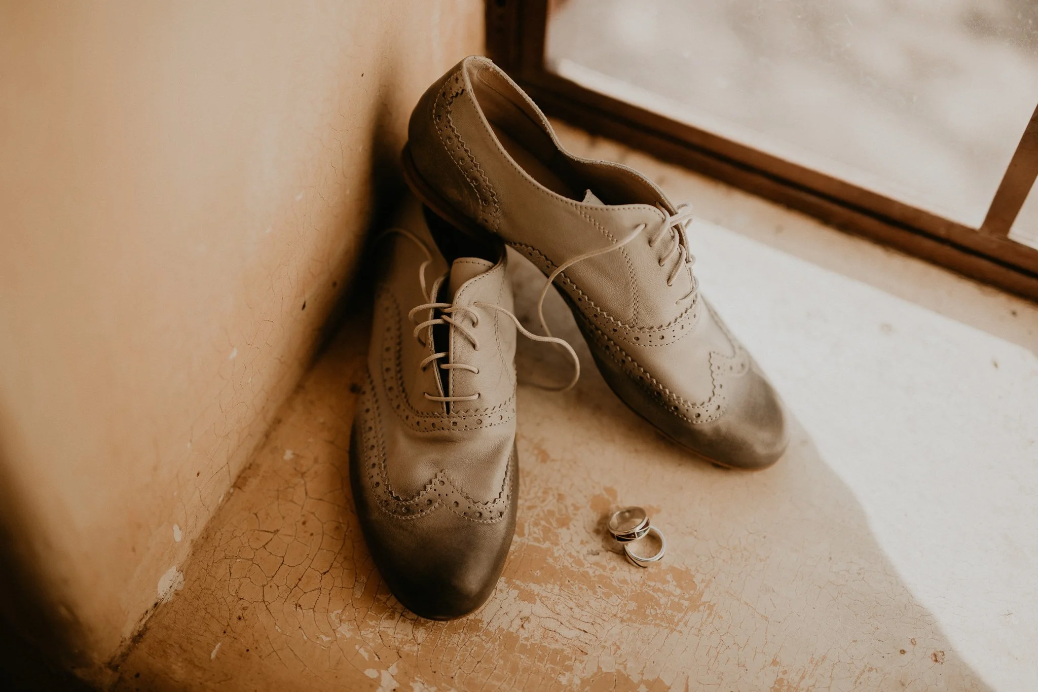A pair of beige and brown dress shoes and two wedding rings on a cracked, beige floor next to a window.