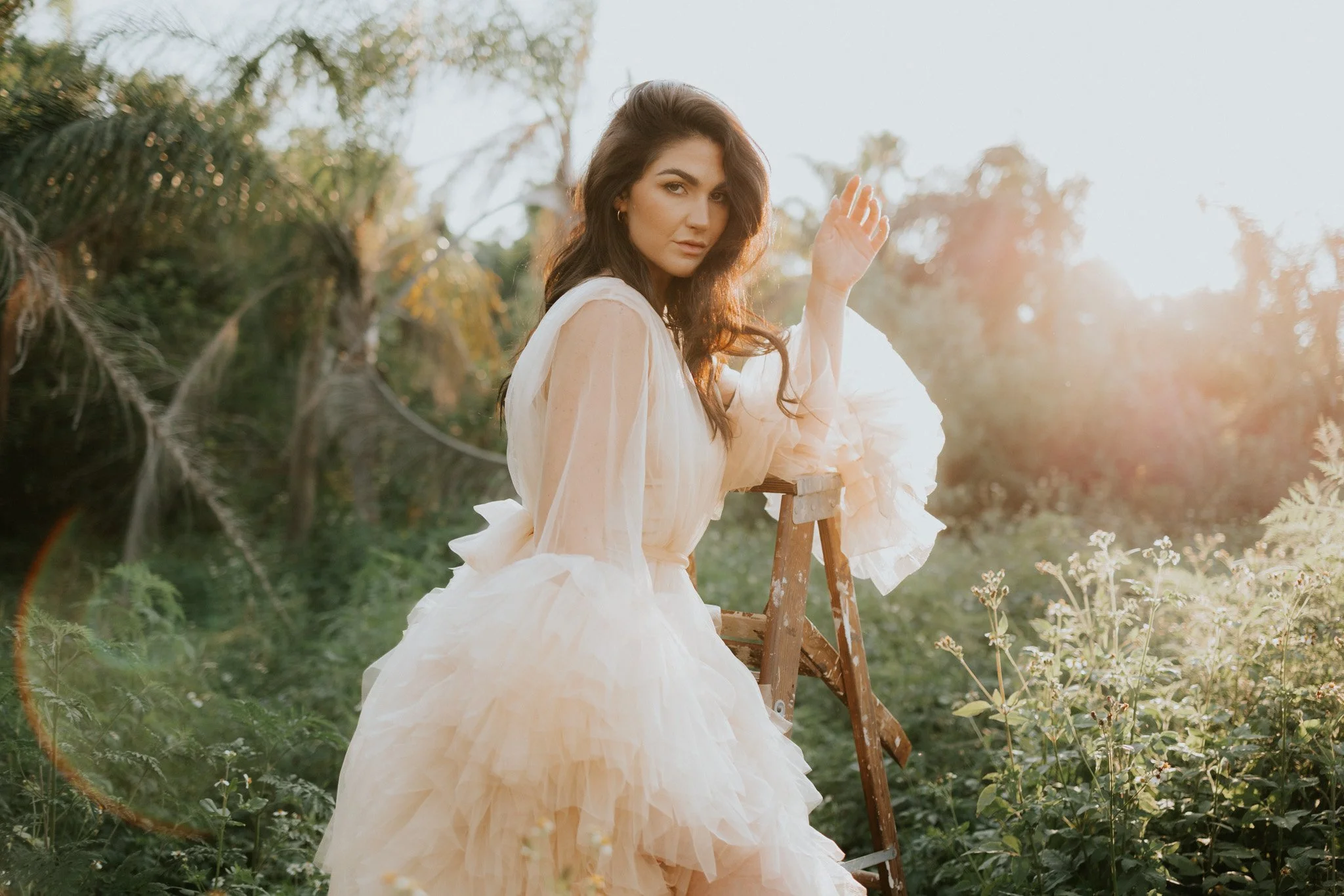 A young woman in a flowing wedding dress sits on a wooden ladder outdoors, surrounded by greenery and plants, with the sunlight shining brightly behind her.