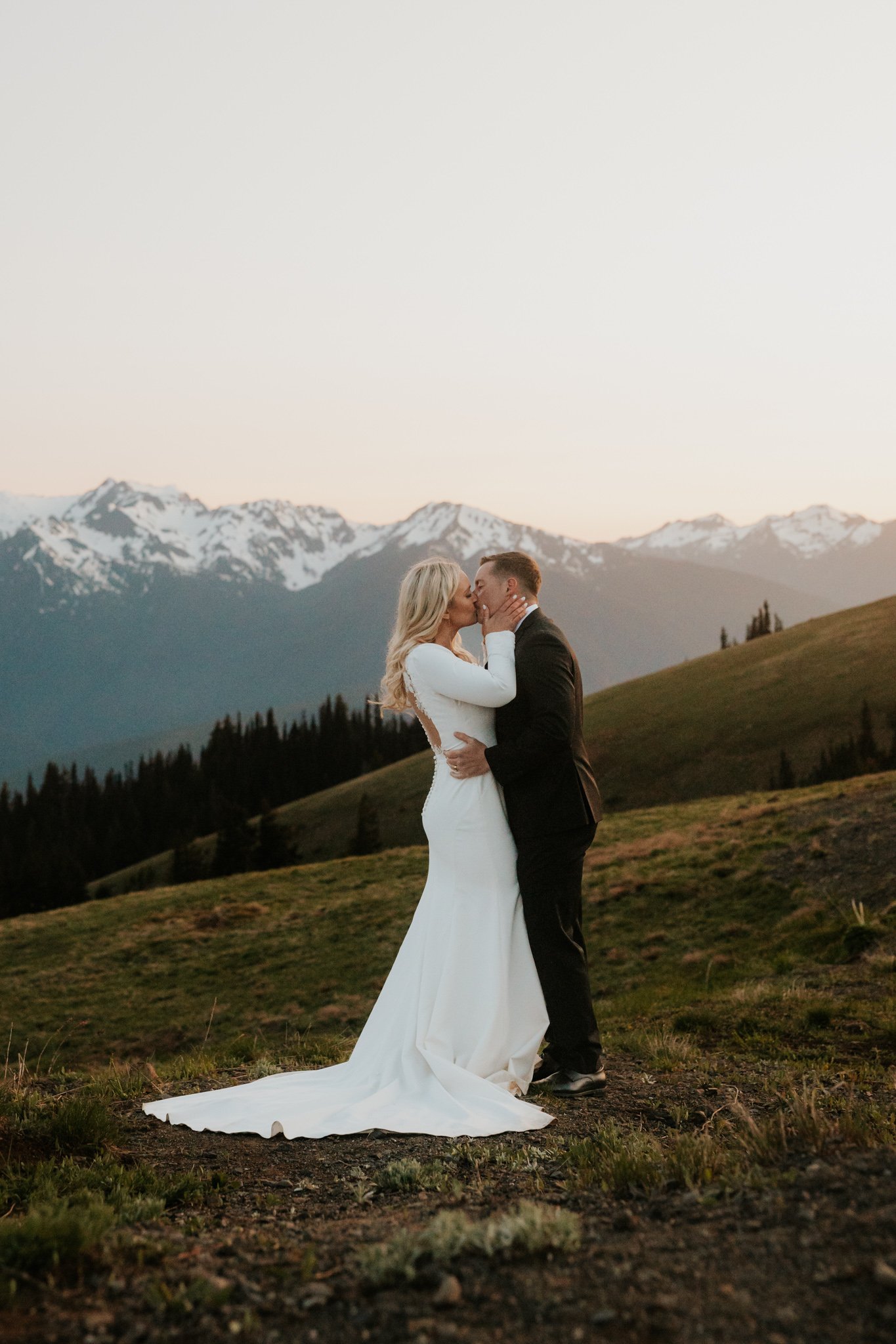 A newlywed couple in wedding attire sharing a kiss on Hurricane Ridge in Olympic National Park at sunset, with snow-capped peaks in the background.