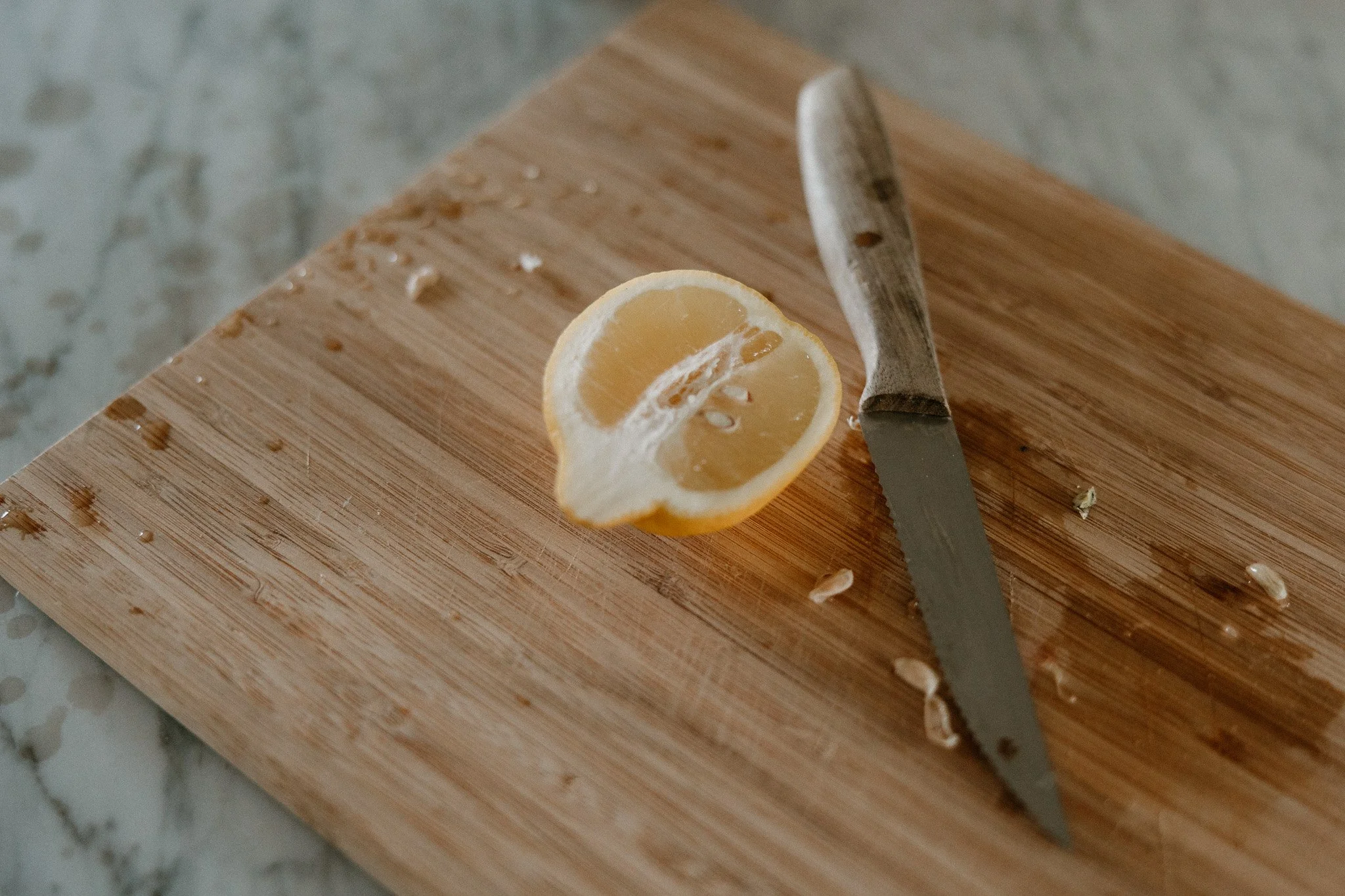 A half lemon on a wooden cutting board with a knife and lemon seeds on the surface.