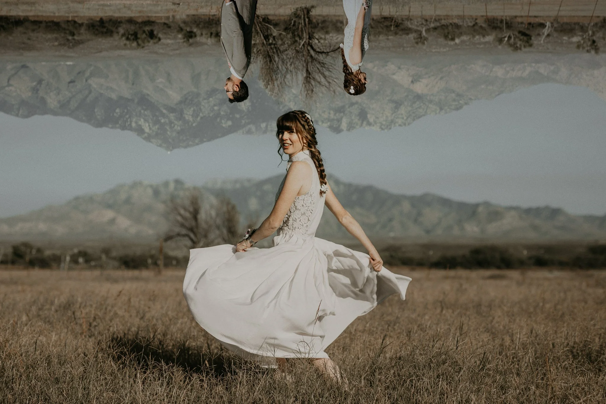 A bride in a white wedding dress standing in a field with mountains and a clear sky in the background, the image is upside down with a reflection effect of the wedding couple.