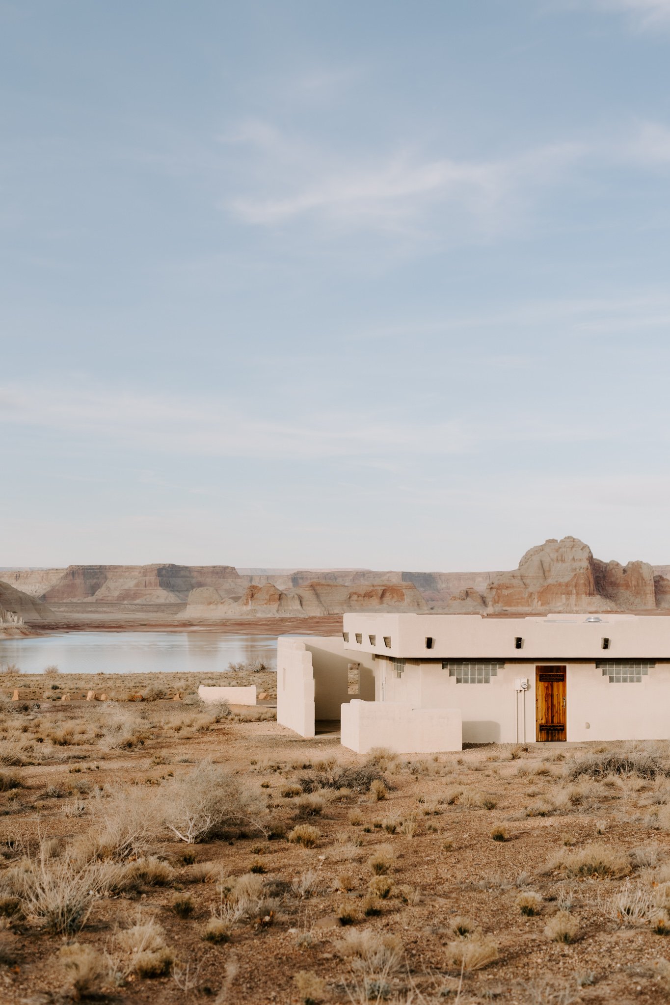A modern white adobe building with a wooden door in a desert landscape with sparse vegetation, a body of water, and mesas in the distance under a partly cloudy sky.