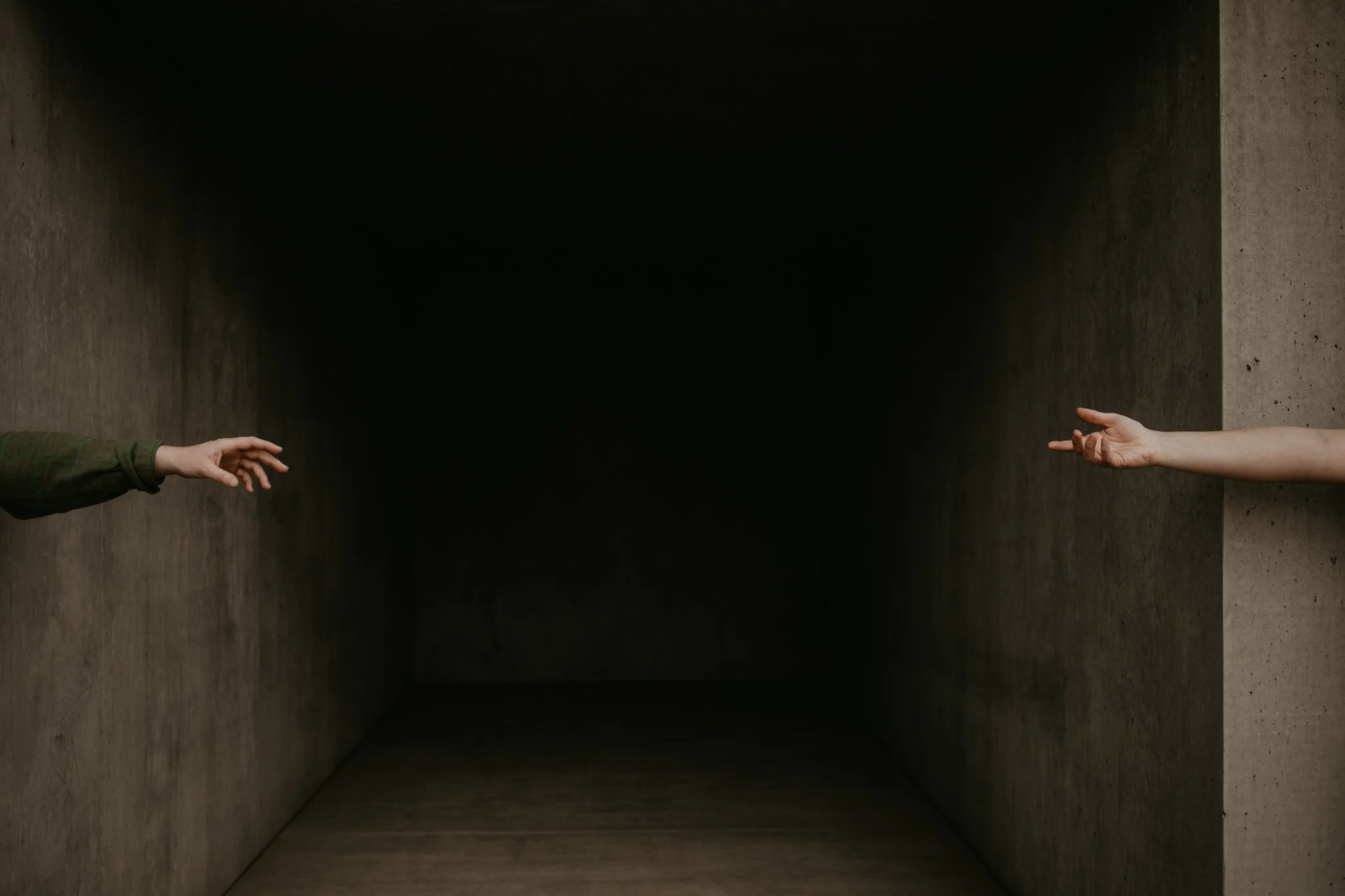 Two hands reaching out from opposite sides of a dark, empty corridor with concrete walls.