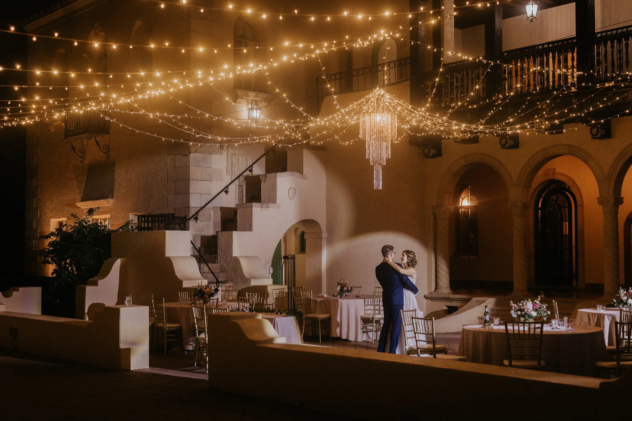 A wedding reception at night with fairy lights and a chandelier, showing a bride and groom dancing in a courtyard decorated with flowers and tables.