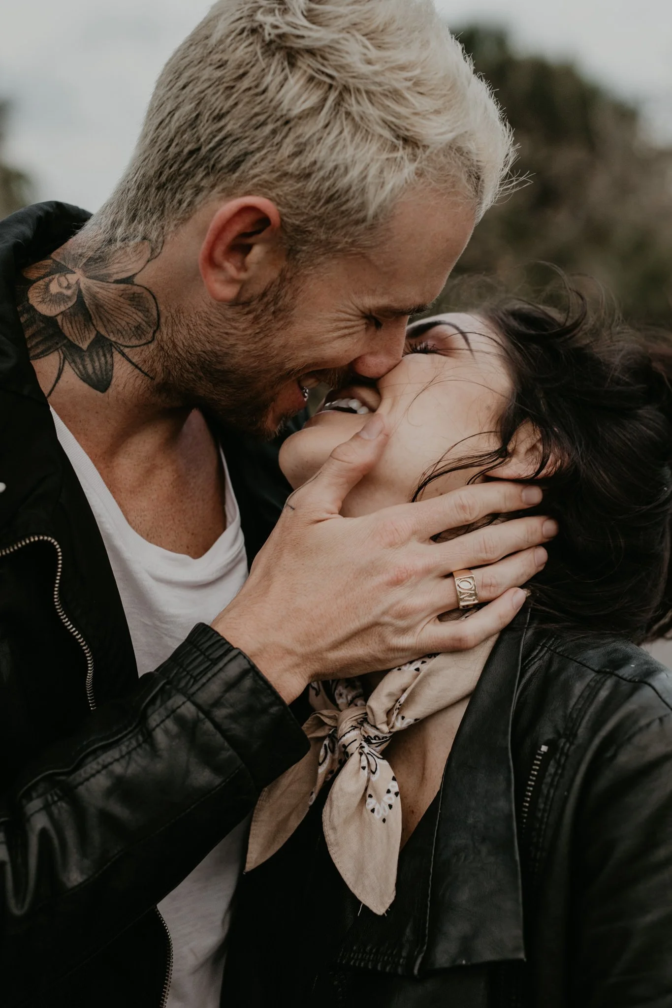A tattooed couple sharing an intimate moment, touching their foreheads and noses together with smiles. The man has a tattoo on his neck, and is wearing a black leather jacket. The woman has dark hair, a bandana, and a black leather jacket.