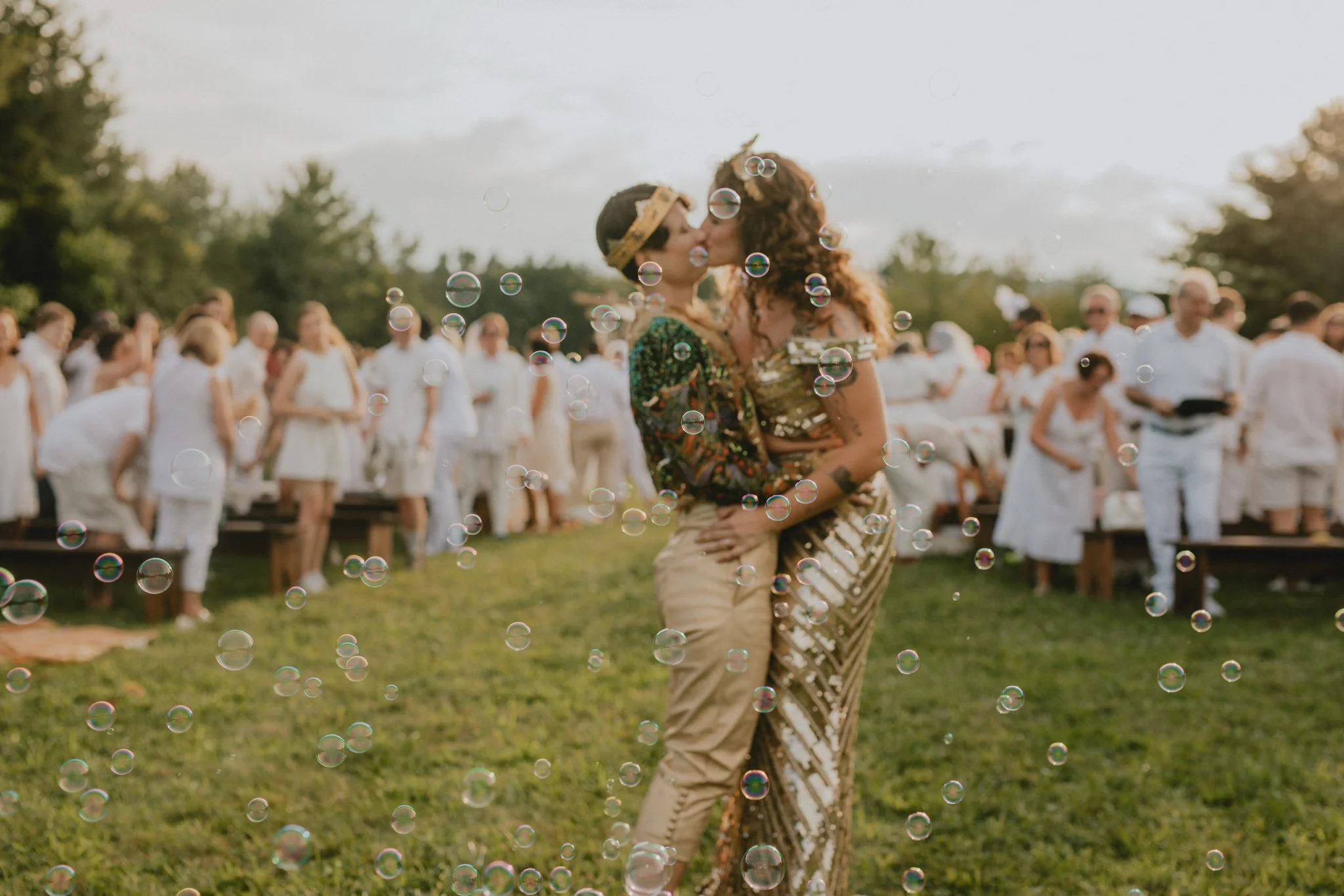 The brides share a kiss, surrounded by bubbles, as their community follows them from their chuppa