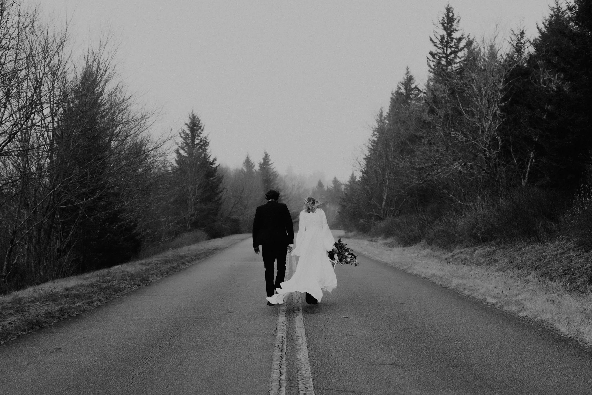 Couple walks a closed portion of the Blue Ridge Parkway on their elopement day, her holding her bouquet by her side