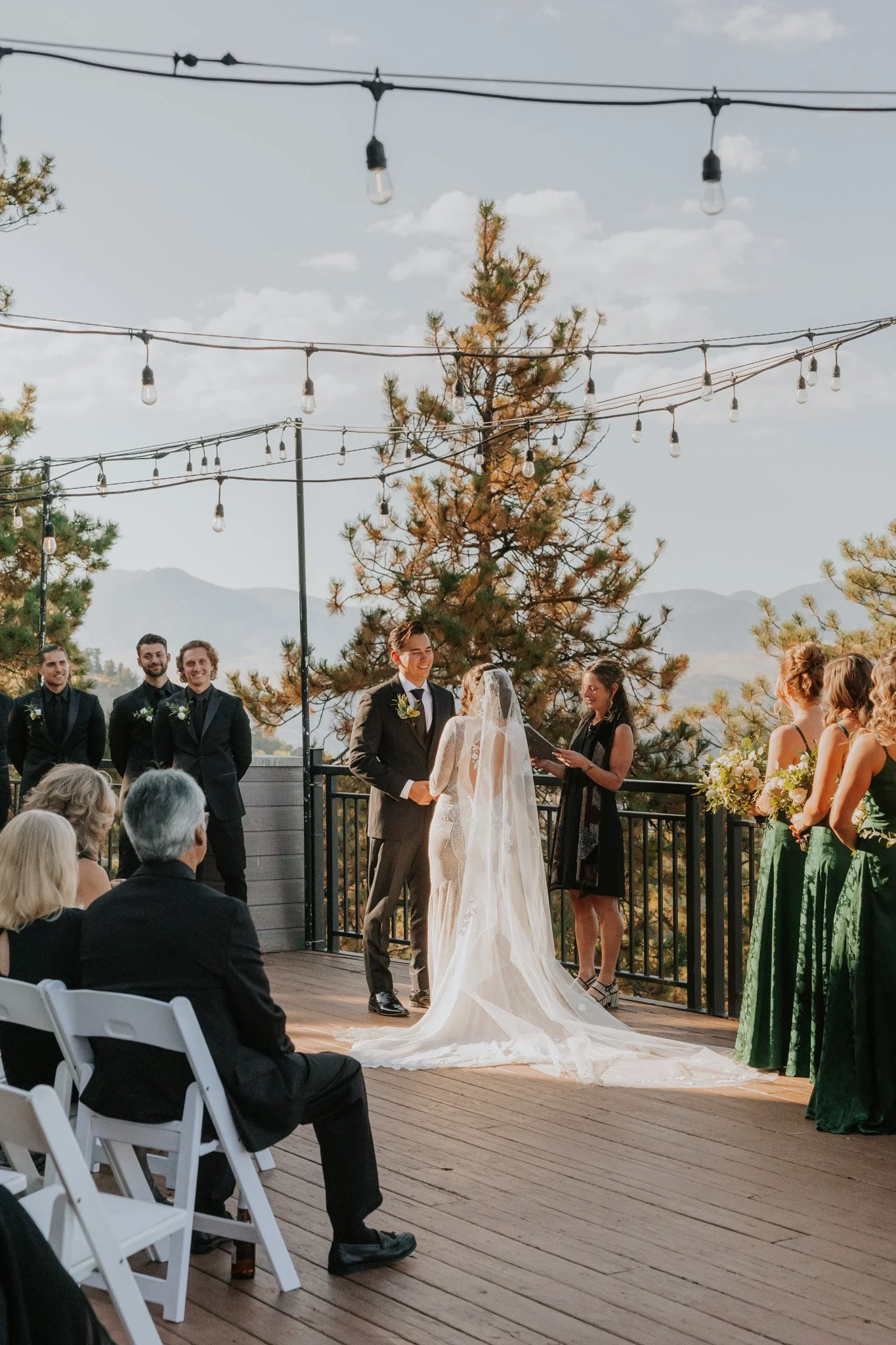 A wedding ceremony taking place outdoors on a wooden deck with mountains and trees in the background. The bride and groom are at the altar, exchanging vows, with the bride in a white wedding gown and veil. Bridesmaids in green dresses and groomsmen i